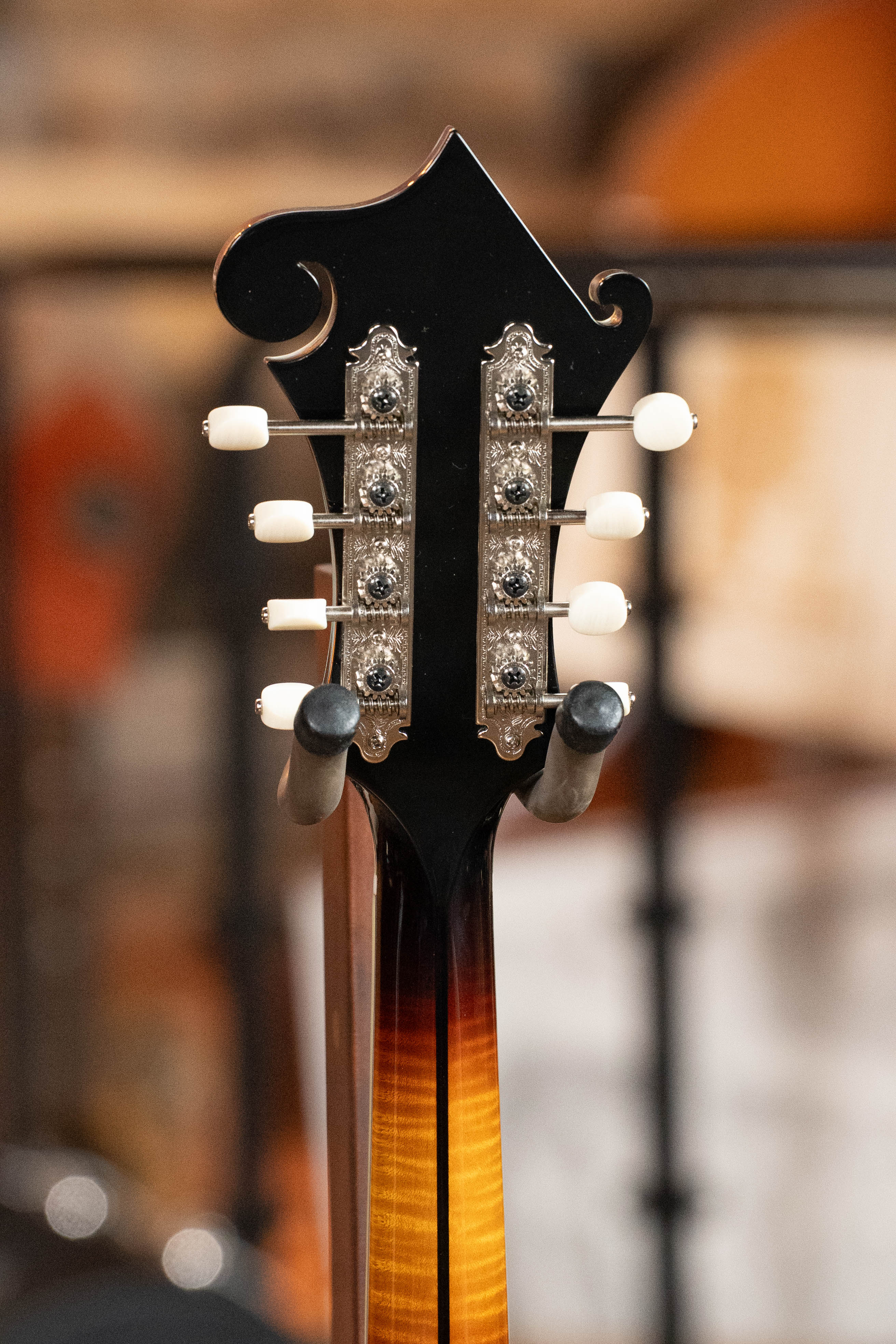 Close-up of the Eastman Guitars MD815-SB Adirondack/Flamed Maple Sunburst F-Style Mandolin #3355 headstock, featuring metal tuning pegs with white knobs and a rich sunburst finish against a blurred background.