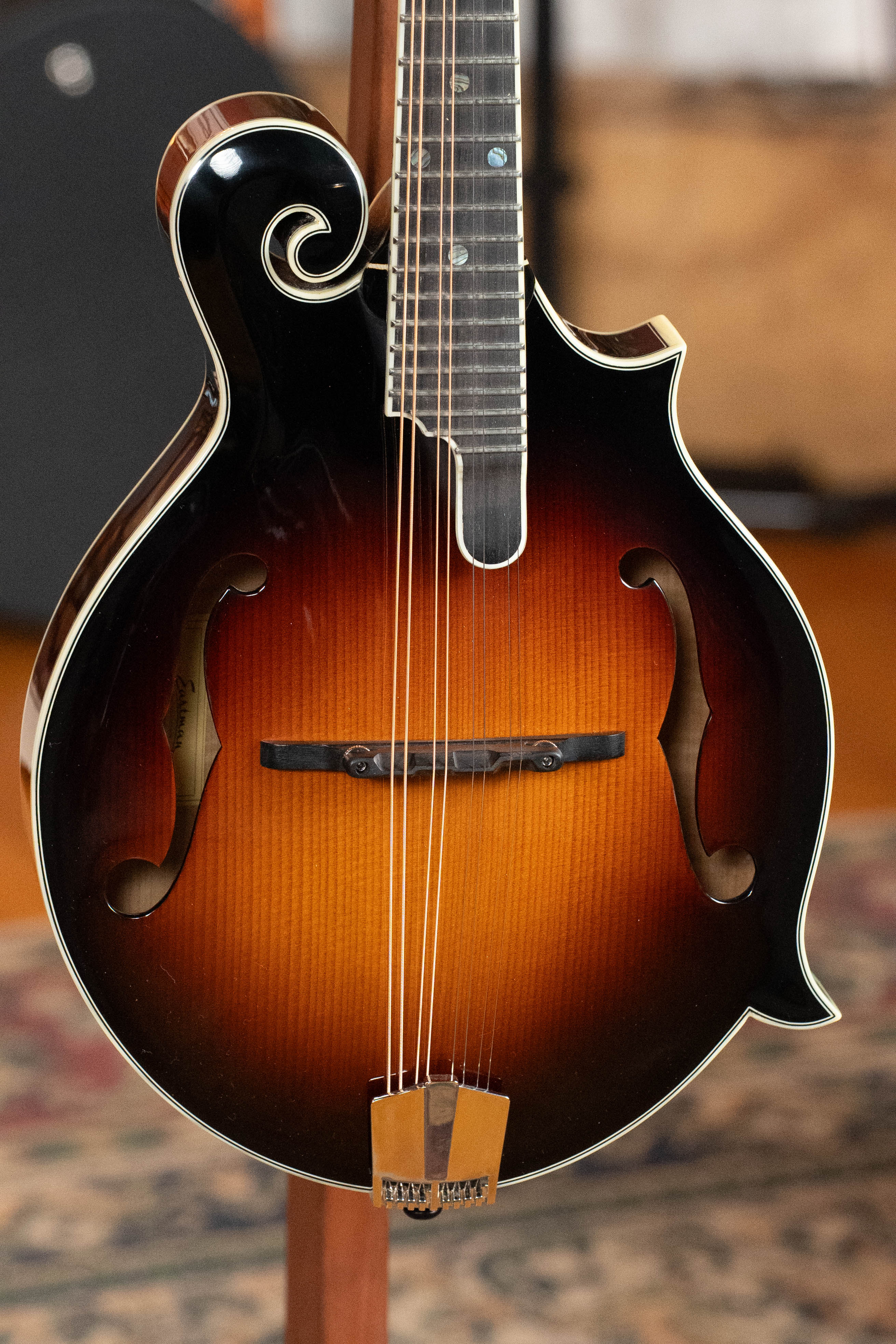 Close-up of an Eastman Guitars MD815-SB Adirondack/Flamed Maple Sunburst F-Style Mandolin #3355, highlighting its scroll, f-holes, and strings. A patterned rug and wood surface appear blurred in the background.