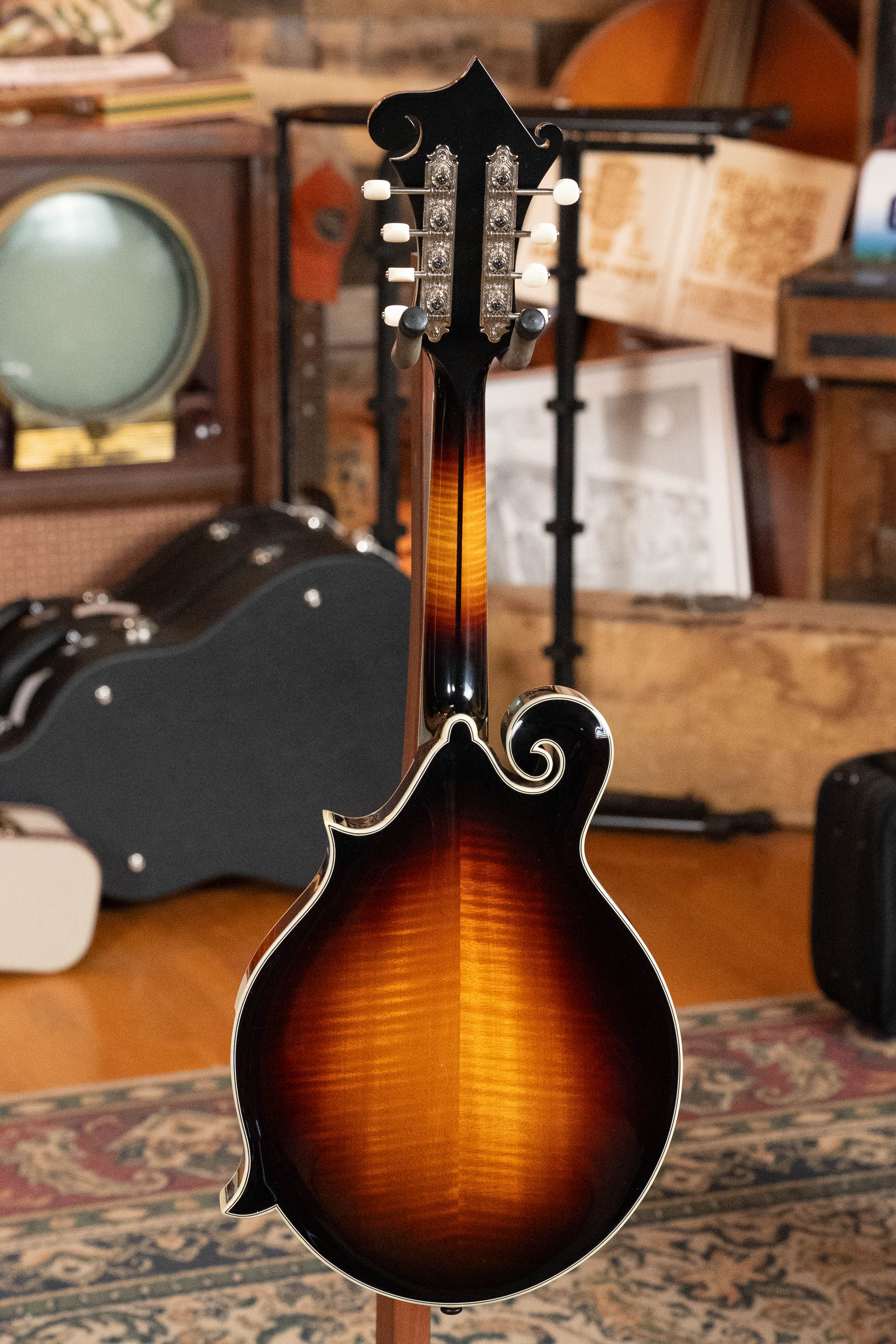 An Eastman Guitars MD815-SB Adirondack/Flamed Maple Sunburst F-Style Mandolin #3355 is shown from the back on a patterned rug, with instruments, cases, a vintage TV, and other objects in the background.
