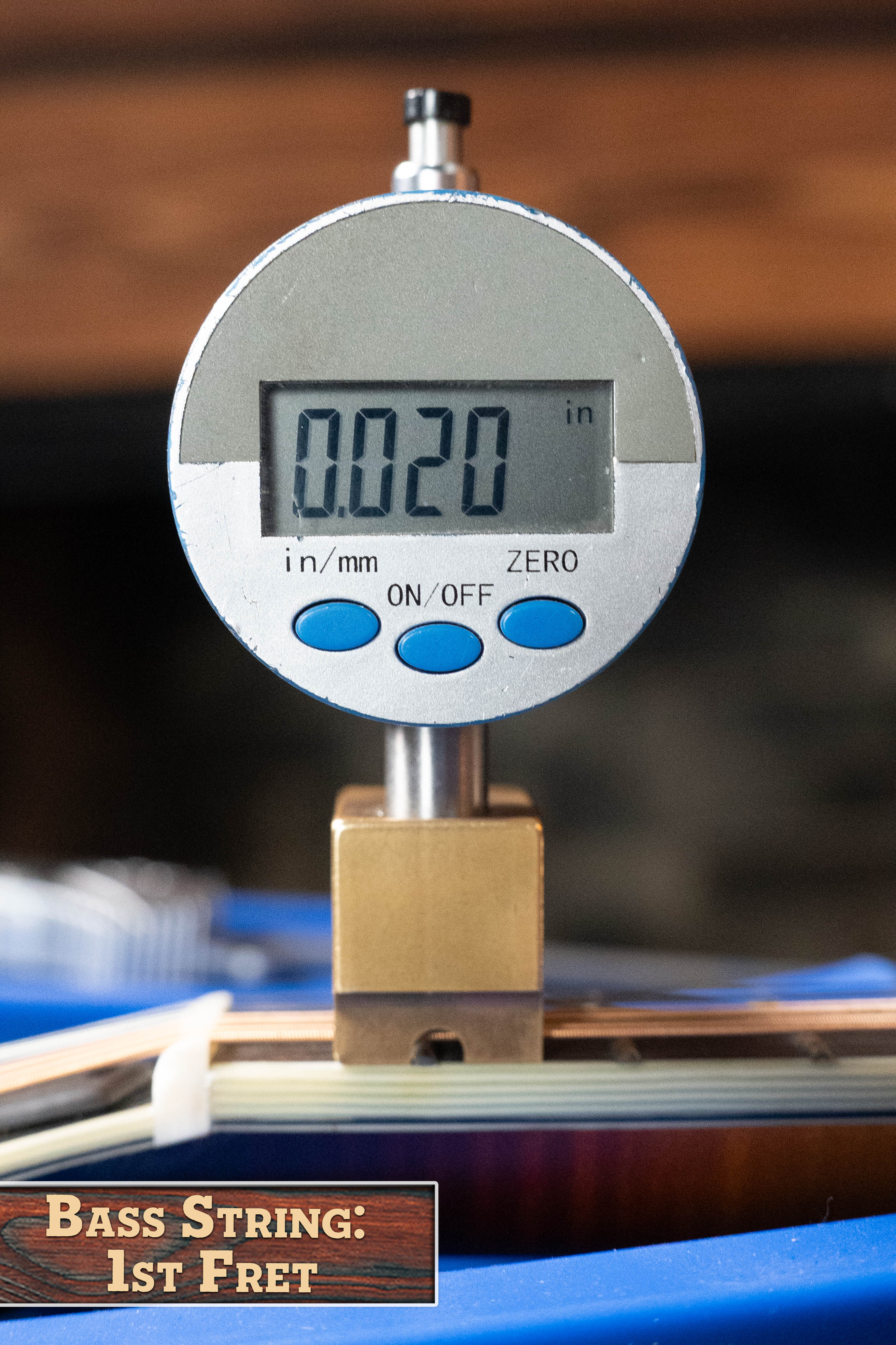 A digital gauge shows 0.020 inches above a bass guitar string at the 1st fret, with a label reading "BASS STRING: 1st FRET." The blurred background recalls the sunburst finish of the Eastman Guitars MD815-SB Adirondack/Flamed Maple Mandolin #3355.