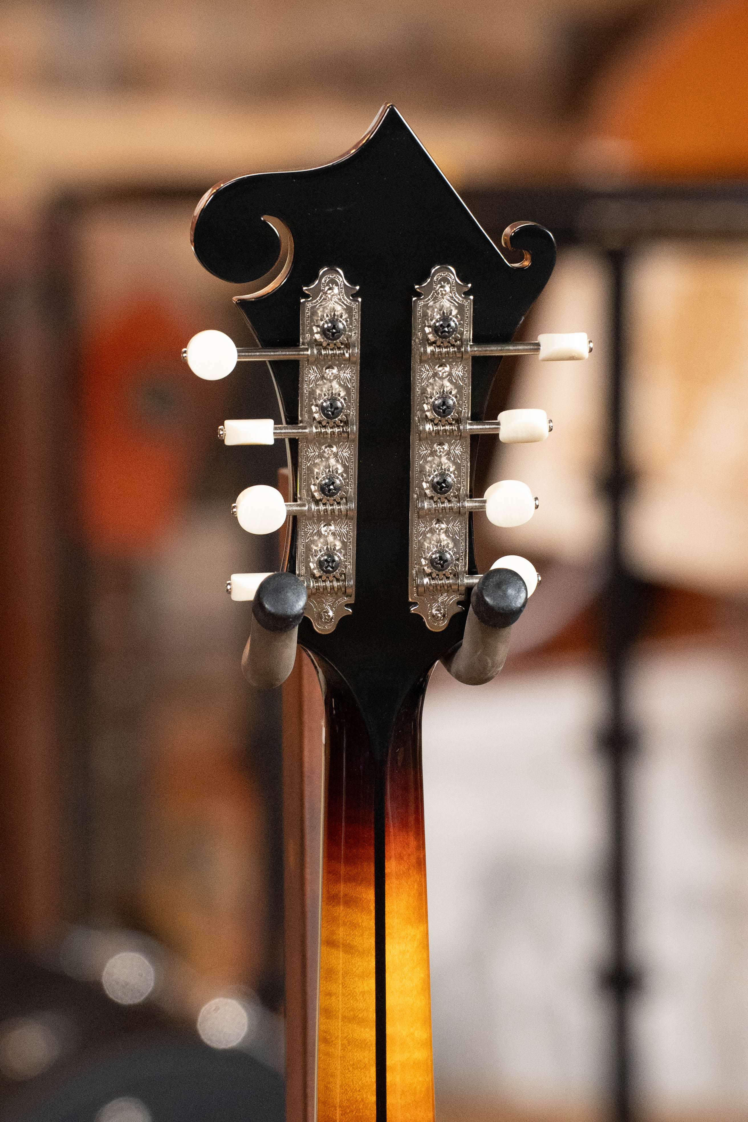 Close-up of the back of an Eastman Guitars MD815-SB Adirondack/Flamed Maple Sunburst F-Style Mandolin #3356 headstock, highlighting tuning pegs and detailed metal gear plates against a blurred background.