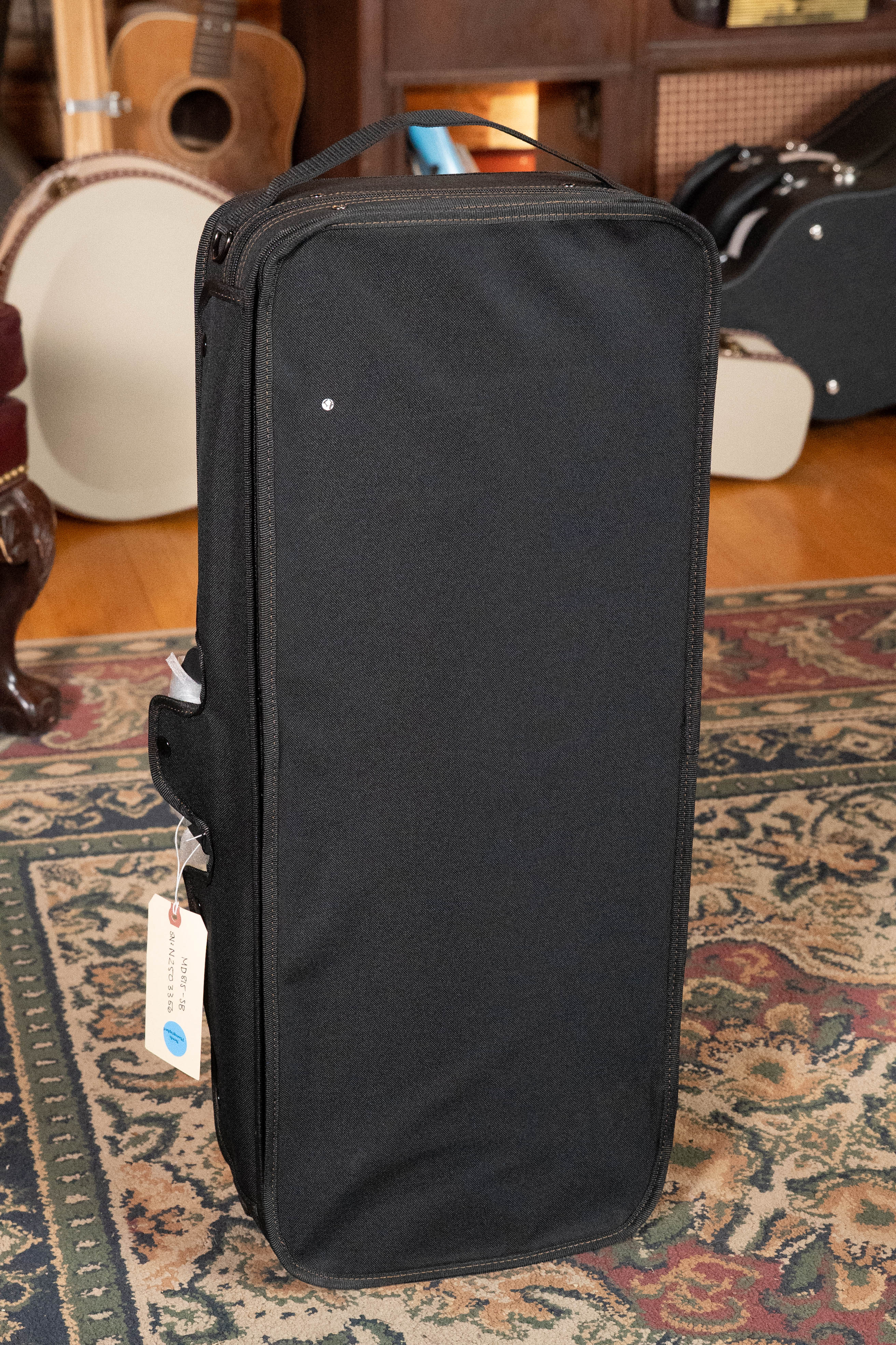 A black rectangular carrying case sits upright on a patterned rug, with an Eastman Guitars MD815-SB Adirondack/Flamed Maple Sunburst F-Style Mandolin #3356 and other instruments visible in the background.