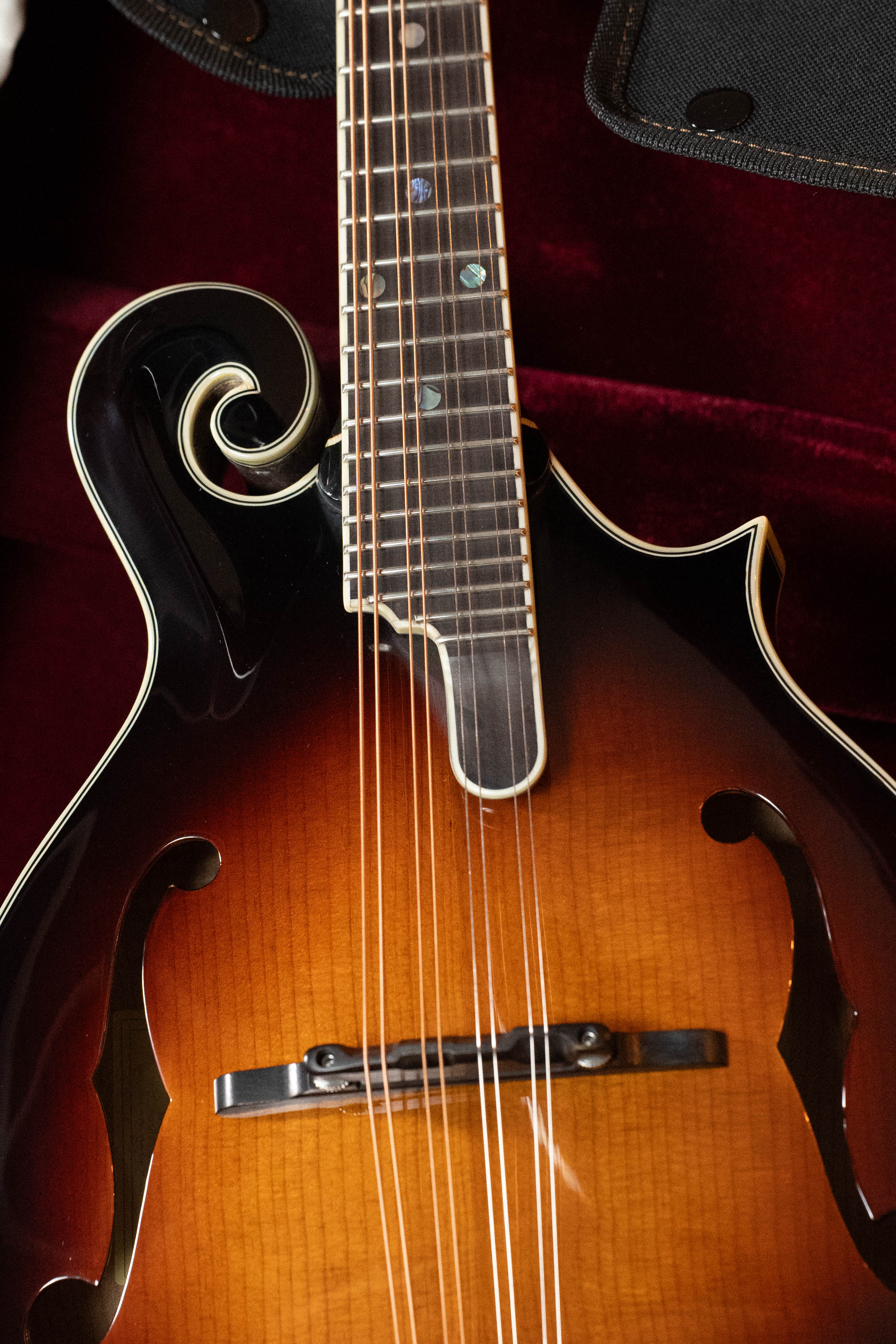 Close-up of an Eastman Guitars MD815-SB Adirondack/Flamed Maple Sunburst F-Style Mandolin #3356, showing decorative scrollwork, f-holes, strings, and fretboard against a dark red velvet background.