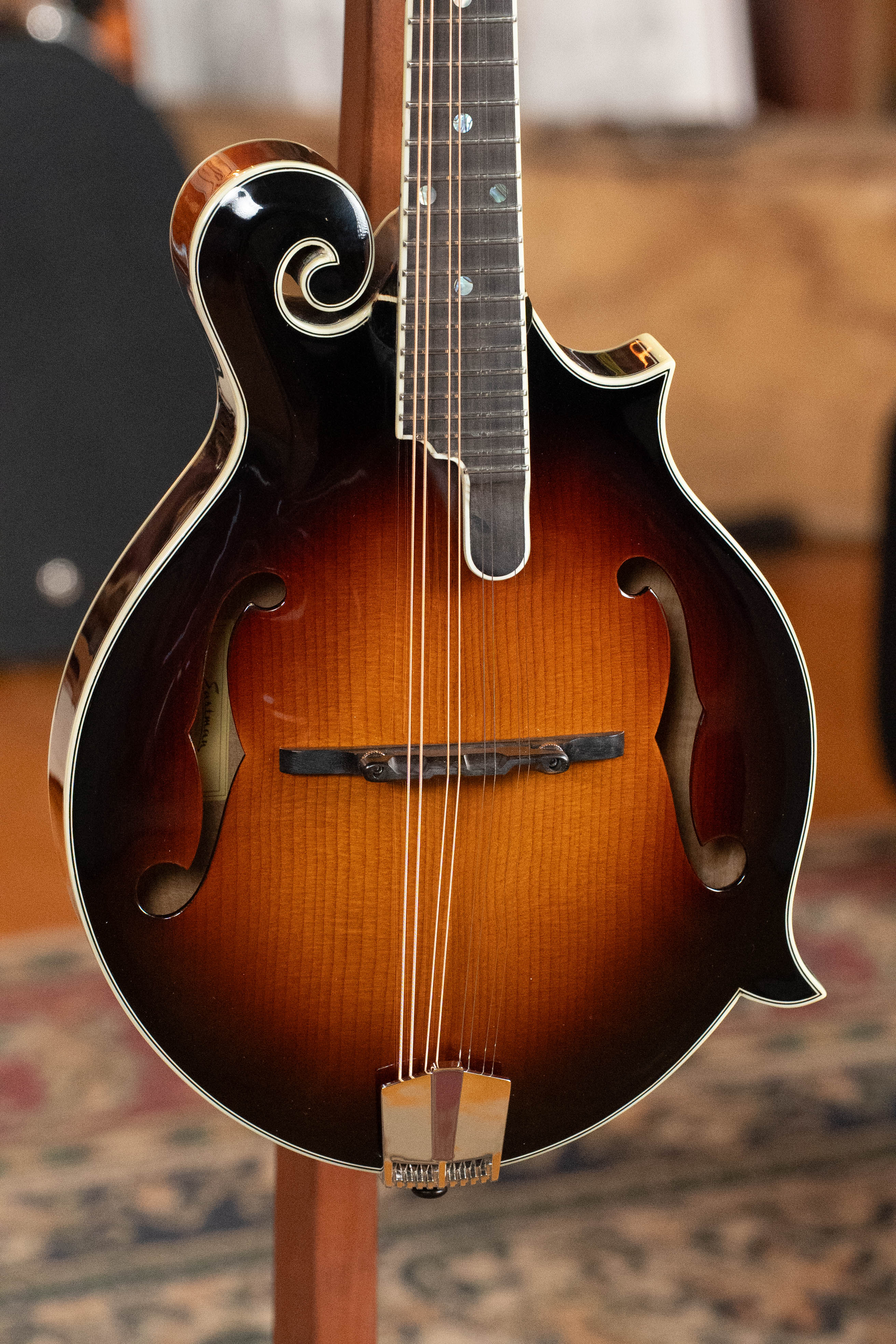 A close-up of an Eastman Guitars MD815-SB Adirondack/Flamed Maple Sunburst F-Style Mandolin #3356, featuring f-holes, an ornate scroll, and gold bridge and tailpiece, shown resting on a stand indoors.