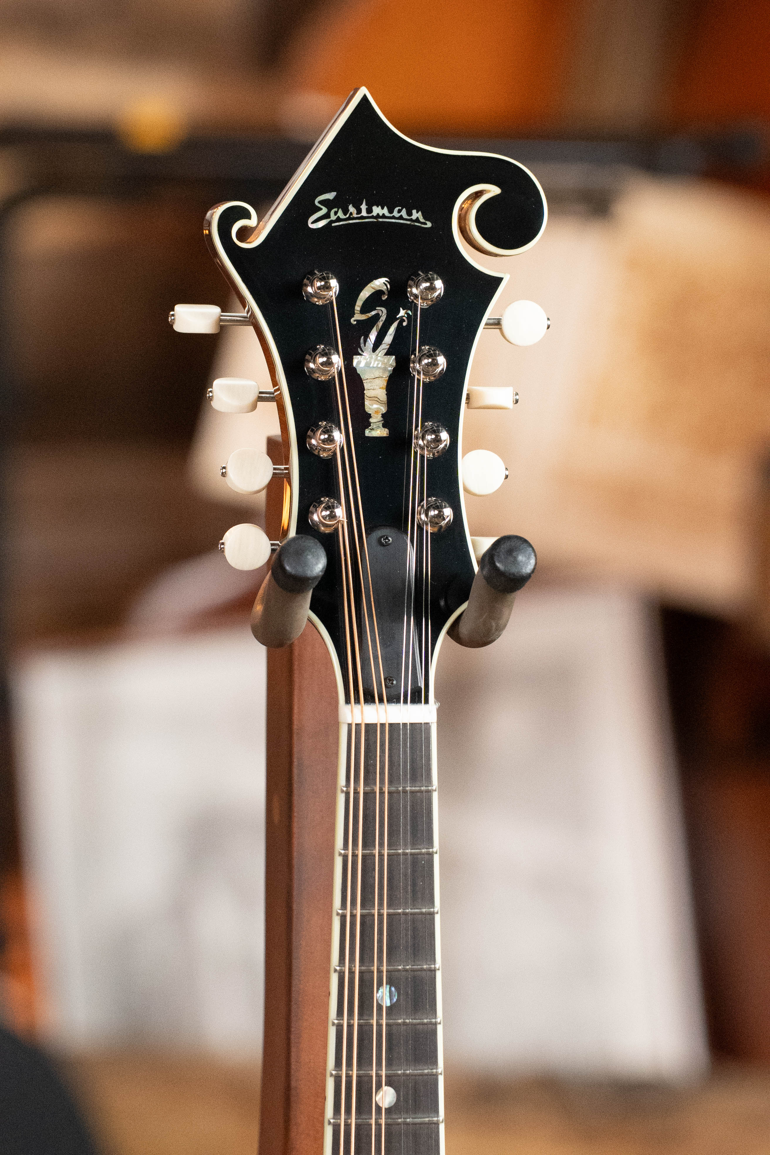 Close-up of the headstock of an Eastman Guitars MD815-SB Adirondack/Flamed Maple Sunburst F-Style Mandolin #3356, featuring white tuning pegs, decorative inlay, and the Eastman logo on black, set against a softly blurred background.