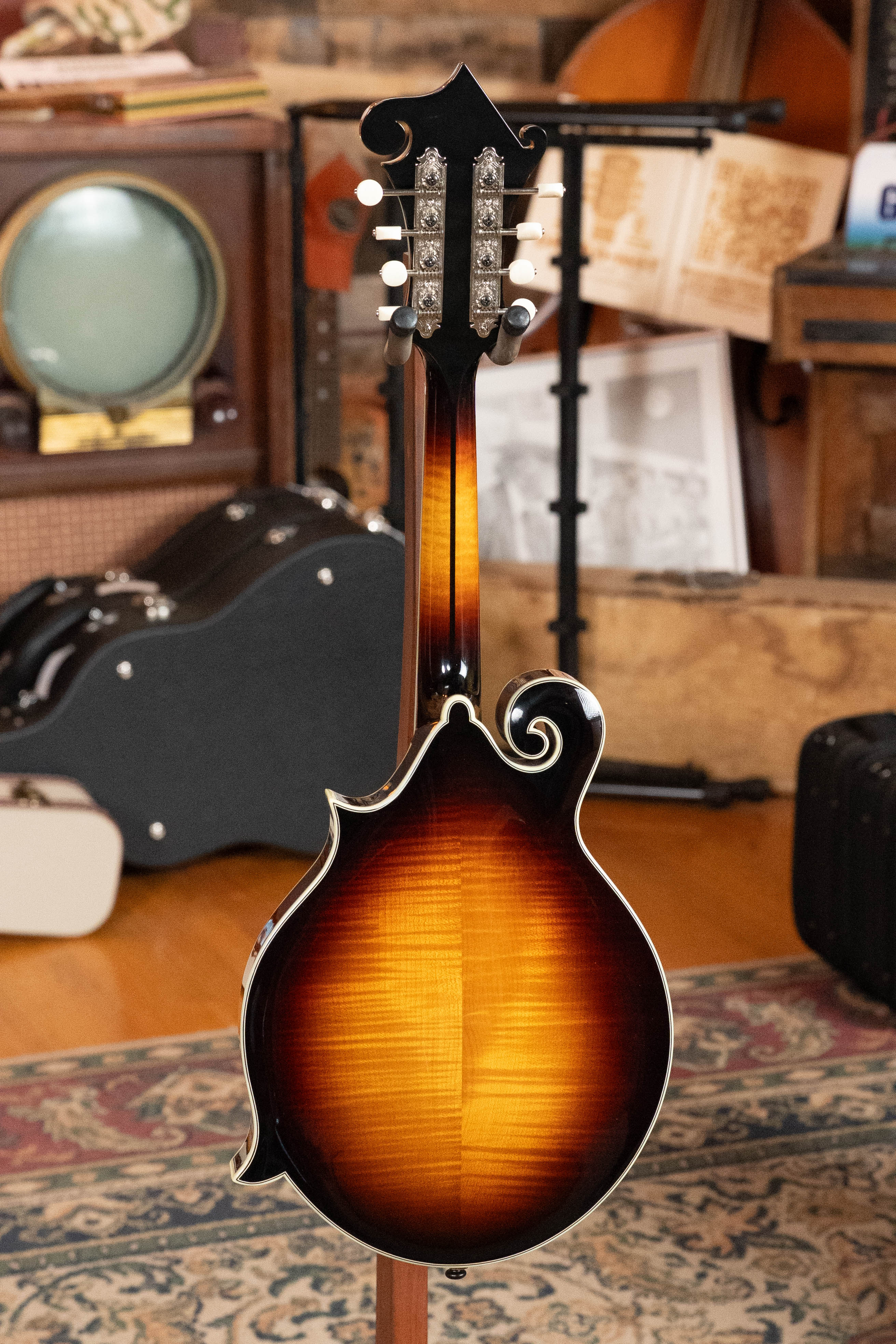 An Eastman Guitars MD815-SB Adirondack/Flamed Maple Sunburst F-Style Mandolin #3356 is shown from the back, standing upright in a cozy room with musical instruments, a vintage TV, and books in the background.