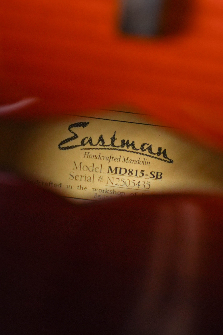 Close-up of a label inside an Eastman Guitars MD815-SB Adirondack/Flamed Maple Sunburst F-Style Mandolin #5435, reading "Eastman Handcrafted Mandolin," with blurred edges of the instrument visible.