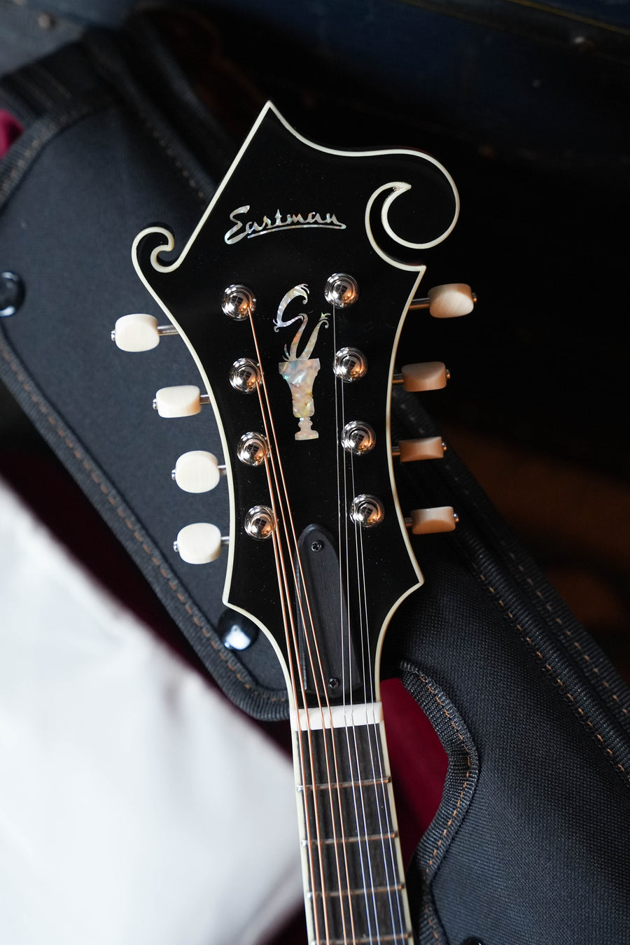 Close-up of the headstock on an Eastman Guitars MD815-SB Adirondack/Flamed Maple Sunburst F-Style Mandolin #5435, showing tuning pegs, strings, the Eastman logo, decorative inlay, and a glimpse of case and background.