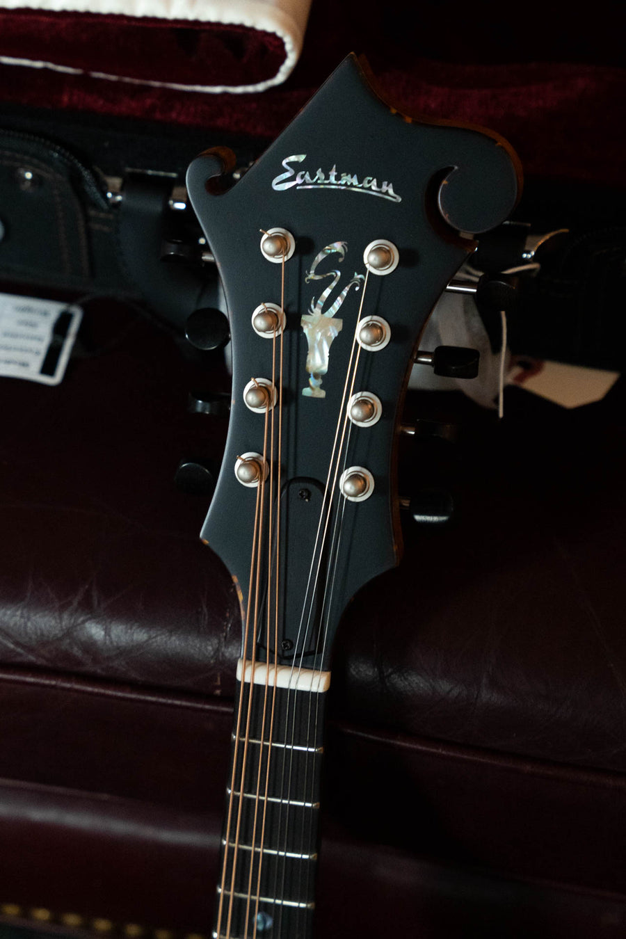 Close-up of the headstock on a black Eastman Strings guitar, featuring tuning pegs, strings, and logo atop dark leather—showcasing craftsmanship like the Eastman MD915-BD Adirondack/Flamed Maple Blonde F-style Mandolin #5819.