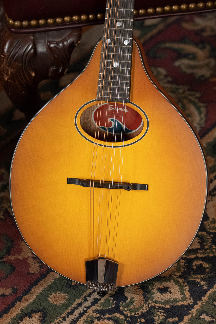 An Eastman Guitars PCH-M104-GB A-Style Mandolin #0056 with a goldburst finish rests on an ornate carpet, teardrop-shaped with an oval soundhole and colorful label, part of a carved wooden chair seen behind it.