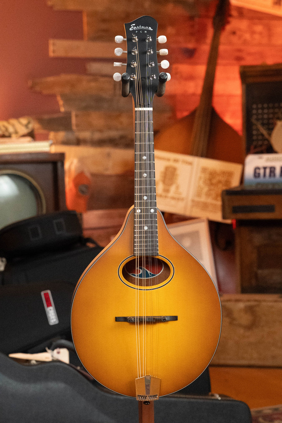 An Eastman Guitars PCH-M104-GB A-Style Mandolin (#0056), featuring a solid spruce top and laminated mahogany back in goldburst finish, sits on a stand in a cozy, wood-paneled room filled with musical instruments and gear.