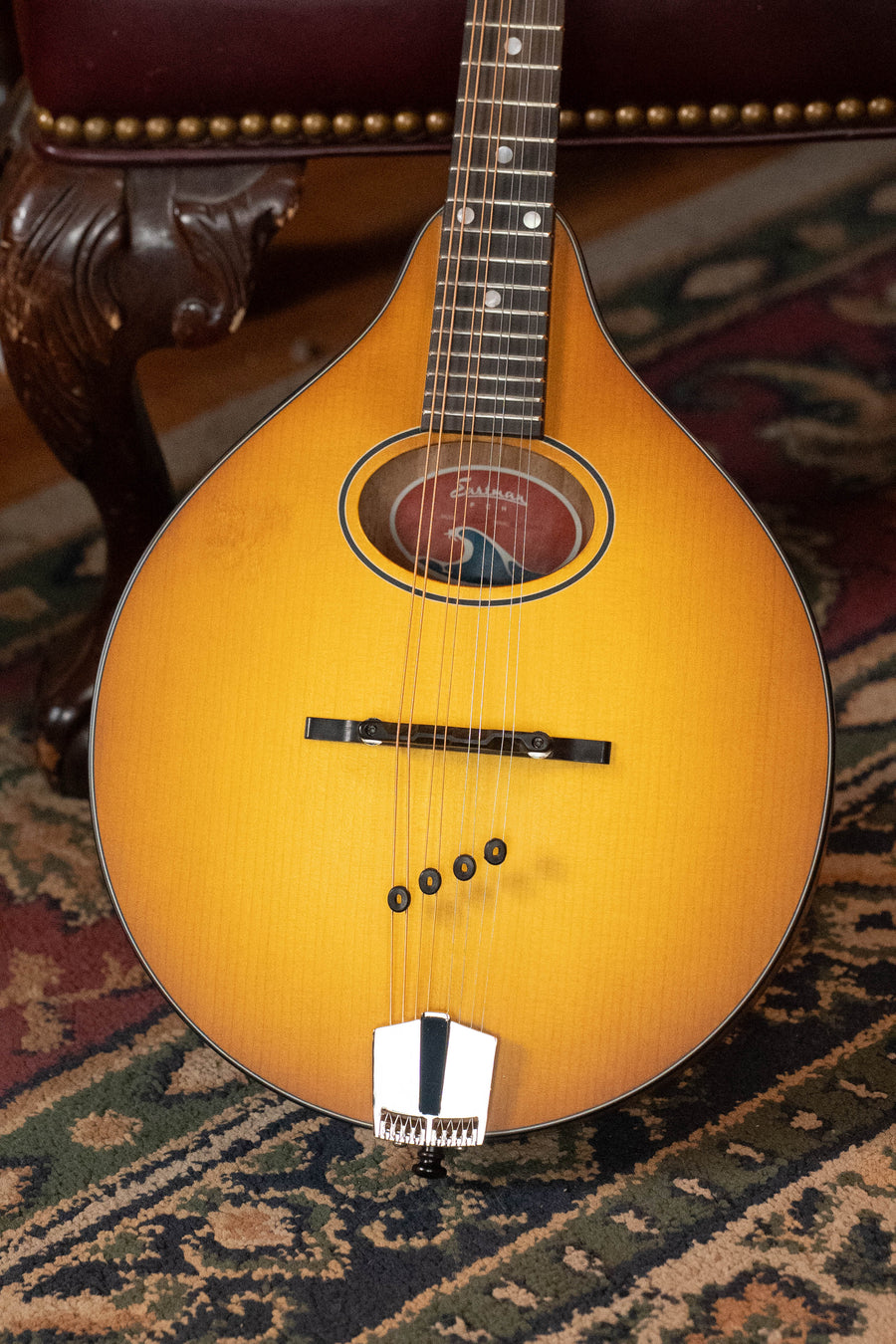A close-up of the Eastman Guitars PCH-M104-GB A-style mandolin (#0063) with 8 strings, solid spruce top, and goldburst finish rests on a patterned rug, with a carved wooden chair in the background—ideal for beginner musicians.
