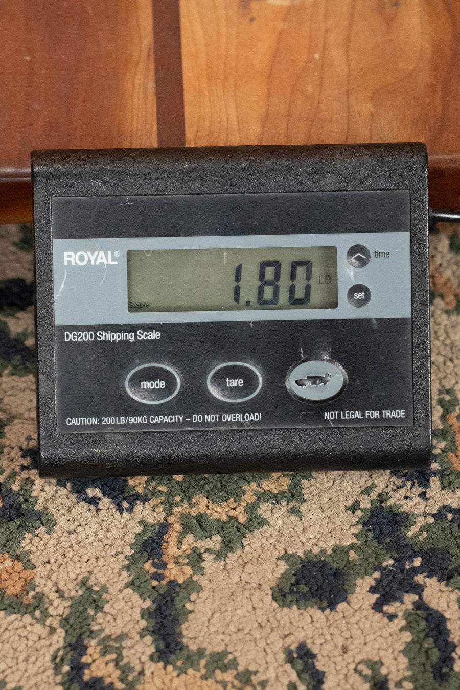 A digital shipping scale on a patterned carpet shows 1.80 pounds while weighing an Eastman Guitars PCH-M104-GB Solid Spruce/Laminated Mahogany Goldburst Flattop A-Style Mandolin #0063. The ROYAL DG200 scale has several buttons and clear labels.