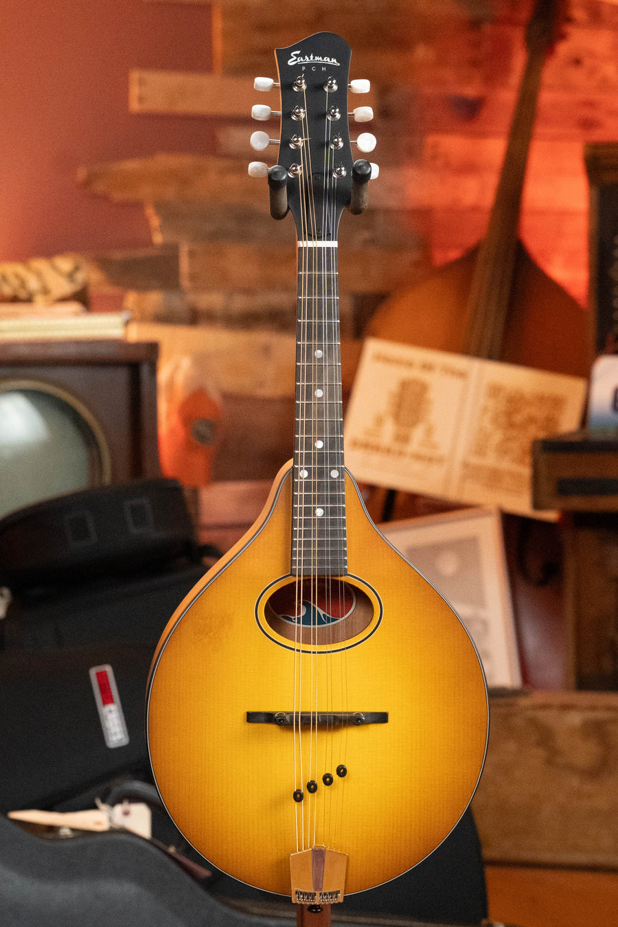 An Eastman Guitars PCH-M104-GB Solid Spruce/Laminated Mahogany Goldburst A-Style Mandolin #0063 is shown upright in a cozy room with wood walls, vintage decor, a music book, and an old TV in the background.