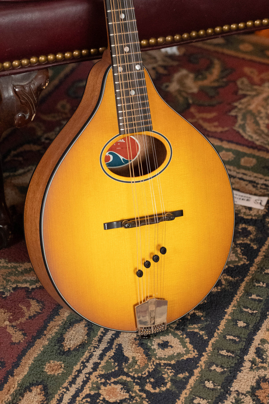 A close-up of the Eastman Guitars PCH-M104-GB A-Style Mandolin #0065, with a Goldburst finish and solid spruce top, rests on a patterned rug—an ideal choice for beginner players seeking quality craftsmanship.