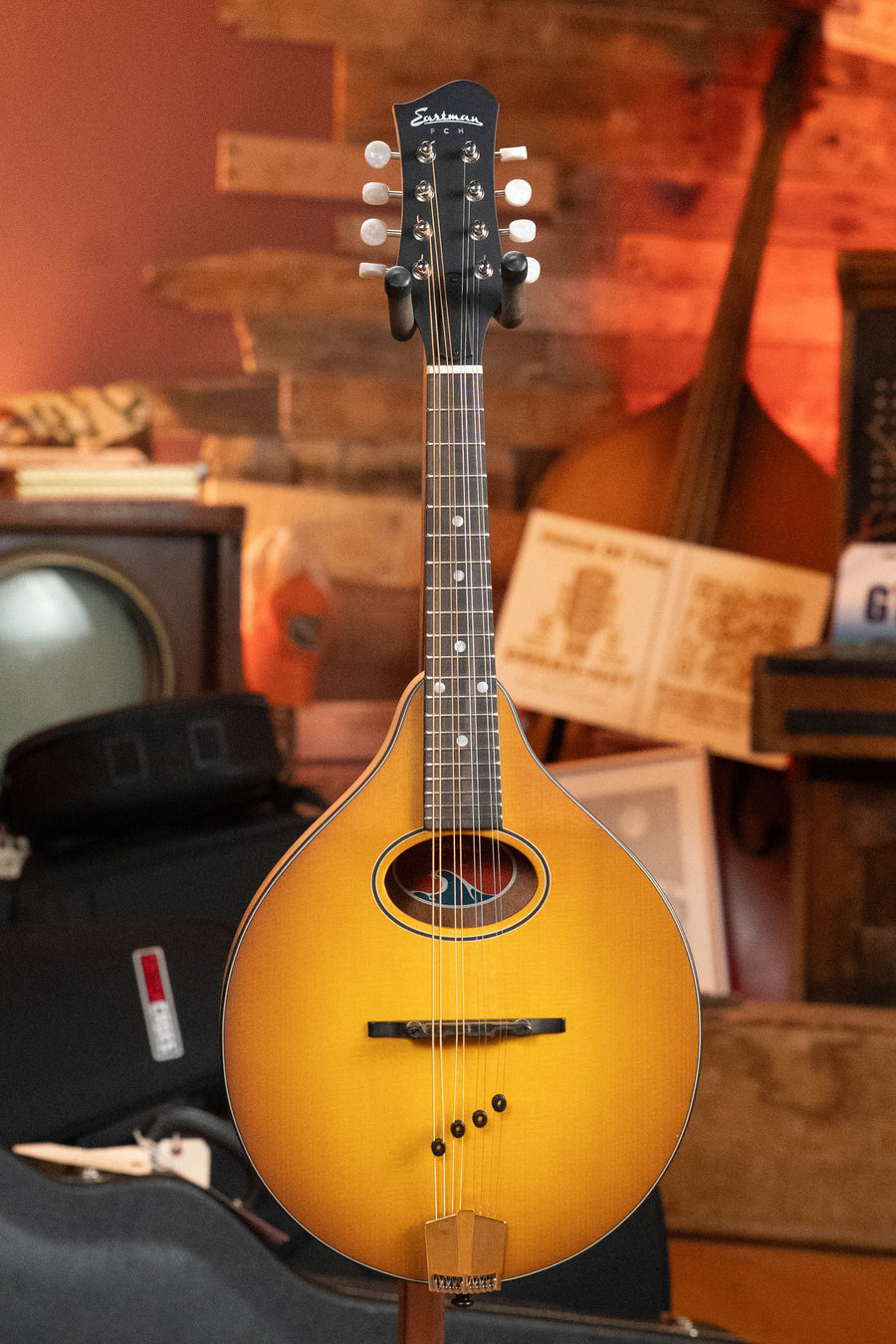 An Eastman Guitars PCH-M104-GB Goldburst A-Style Mandolin with a solid spruce top and laminated mahogany body stands upright in a cozy room featuring wooden walls, vintage equipment, and musical instruments.