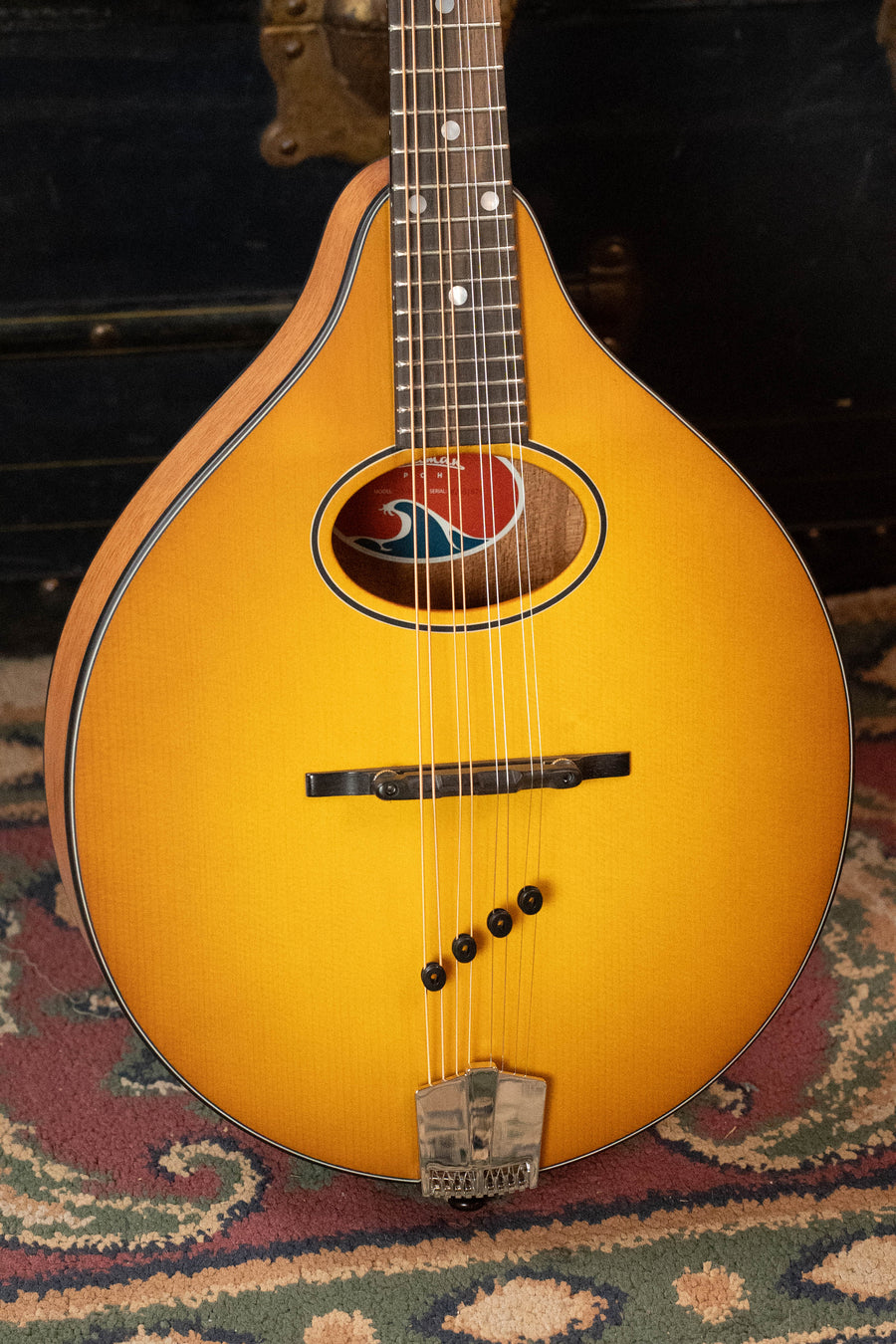 An Eastman Guitars PCH-M104-GB Mandolin (#1876) with a goldburst flattop A-style body, solid spruce top, laminated mahogany back/sides rests on a patterned rug, making it an ideal beginner mandolin. Part of a dark chest is visible in the background.