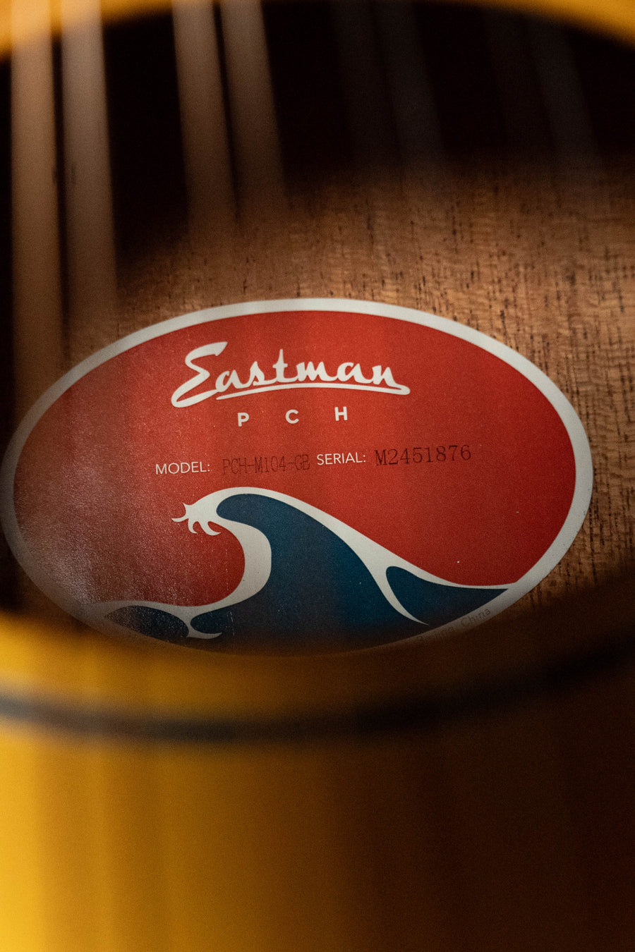 Close-up of a mandolin’s soundhole, showing the Eastman Guitars label inside. The label says PCH-M104-GB with model and serial #1876, features a blue wave graphic, and sits on visible wood grain of solid spruce/laminated mahogany.