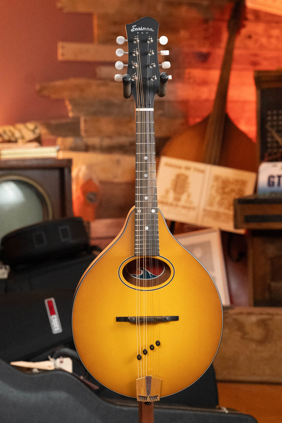 An Eastman Guitars PCH-M104-GB Solid Spruce/Laminated Mahogany Goldburst Flattop A-Style Mandolin (#1876) stands upright in a warmly lit, wood-paneled room with other musical instruments in the background.