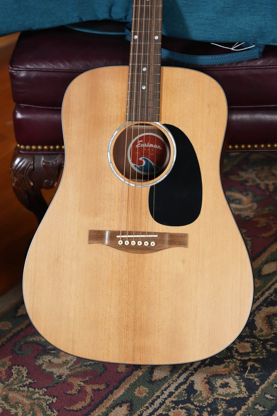 A close-up of an Eastman Guitars PCH1-D #1819 dreadnought acoustic with a solid Sitka spruce top and laminated sapele back/sides, black pickguard, and six strings, resting on a patterned rug beside a red cushioned chair.