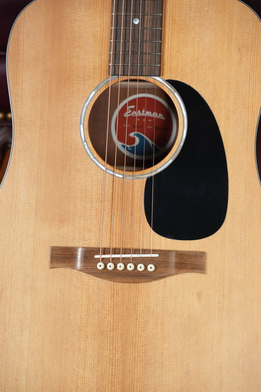 Close-up of an Eastman Guitars PCH1-D Solid Sitka/Laminated Sapele Dreadnought Acoustic Guitar #1819, featuring six strings, a solid Sitka spruce top, wooden bridge with white pins, black pickguard, and round sound hole with red and white label.
