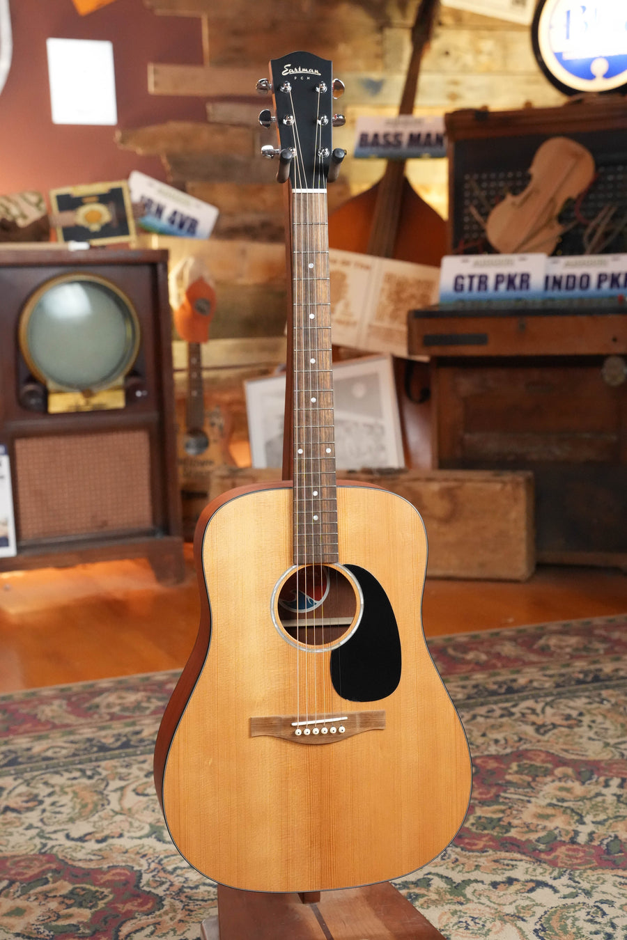 An Eastman Guitars PCH1-D Solid Sitka/Laminated Sapele Dreadnought Acoustic Guitar #1819 stands upright on a patterned rug in a cozy room with vintage decor, featuring an old TV, wooden crates, and musical signs in the background.