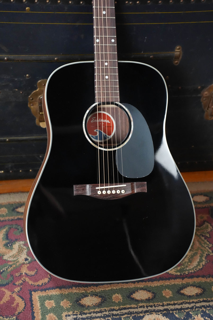 The Eastman Guitars PCH2-D-BK Solid Sitka/Laminated Rosewood Black Top Dreadnought Acoustic Guitar #7988 stands upright on a patterned rug, featuring a glossy black finish, pickguard, visible strings, and a solid Sitka spruce top.