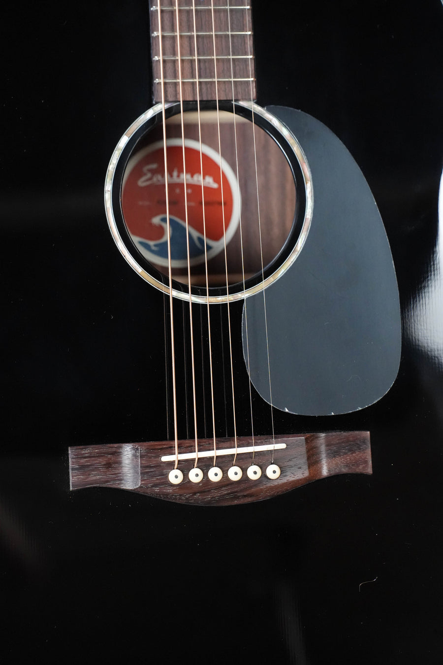 Close-up of an Eastman Guitars PCH2-D-BK Dreadnought #7988, showing its solid Sitka spruce top, black finish, sound hole with red label, pickguard, strings, and part of the bridge.