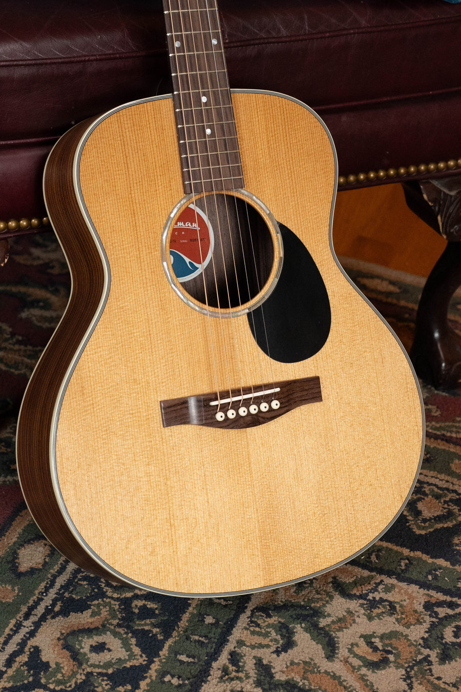 An Eastman Guitars PCH2-TG Thermo-Cured Solid Sitka/Laminated Rosewood Travel Acoustic Guitar (#7477) leans against a maroon leather sofa on a patterned rug.