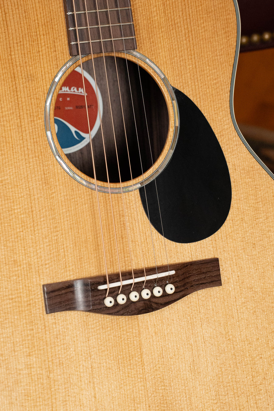 Close-up of the Eastman PCH2-TG Travel Acoustic Guitar by Eastman Guitars, showing its thermo-cured solid Sitka spruce top, sound hole, strings, wooden bridge with six white pins, black pickguard, and light wood body.