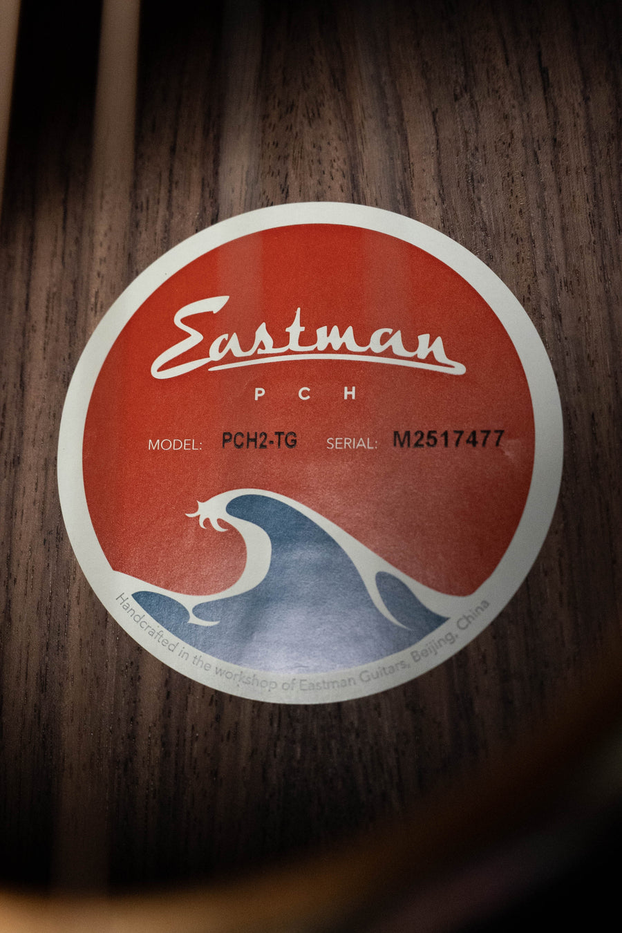 A close-up of an Eastman GCH2-TG Thermo-Cured Solid Sitka/Laminated Rosewood Travel Acoustic Guitar #7477 label, showing model and serial number, featuring a wave graphic—handcrafted by Eastman Guitars in Beijing, China.