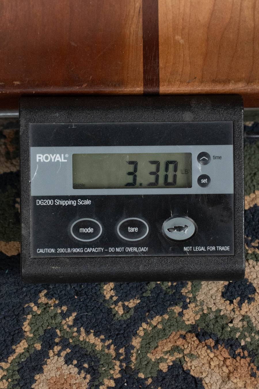 A digital shipping scale on a patterned carpet displays an Eastman Guitars PCH2-TG Thermo-Cured Travel Acoustic Guitar #7477 weighing 3.30 pounds. The scale’s panel features several buttons and warnings beside the lightweight Eastman instrument.