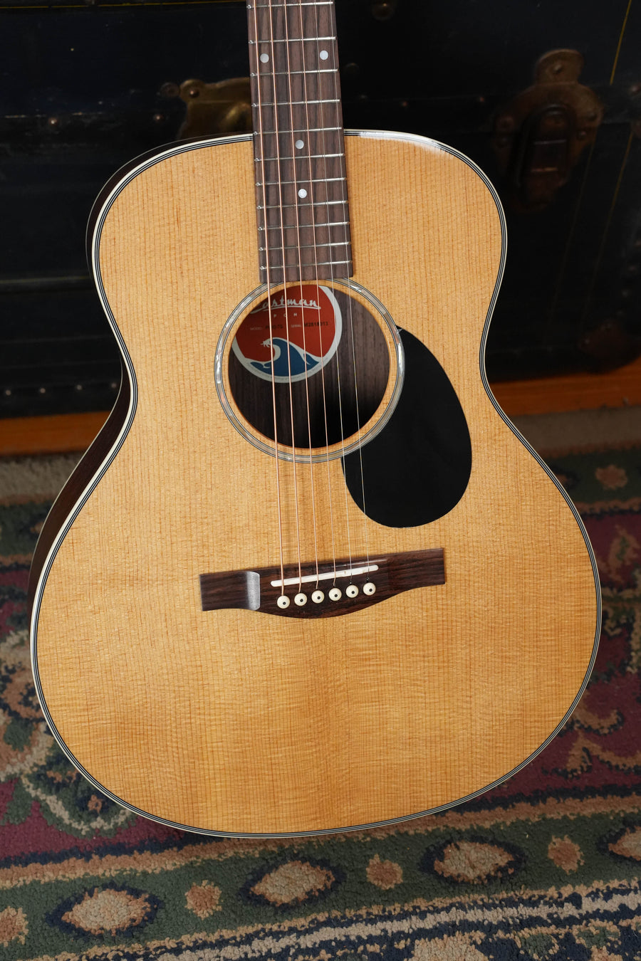 The Eastman Guitars PCH2-TG Travel Acoustic Guitar #8013, featuring a thermo-cured solid Sitka top and laminated rosewood, rests on a patterned rug, displaying its strings, soundhole, pickguard, and part of the fretboard.
