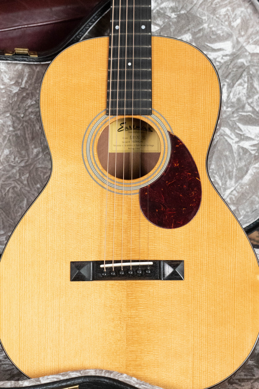 A close-up of the Eastman Guitars E10OO-TC Thermo-Cure Adirondack/Mahogany Acoustic Guitar #4295 highlights its Adirondack spruce top, visible strings, dark pickguard, and part of a case—echoing classic Pre-War parlor guitar style.