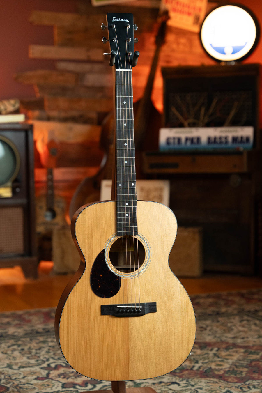 An Eastman Guitars E1OML All Solid Sitka/Sapele Left-Handed Acoustic Guitar #8025 with a light wood body and dark pickguard stands upright in a warmly lit room featuring vintage decor, including retro TV, wooden furniture, and musical gear.