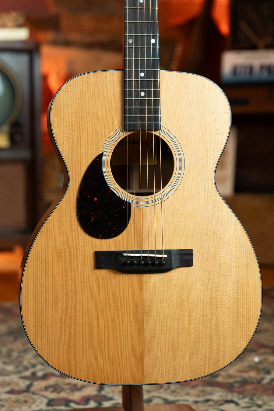 A close-up of the Eastman Guitars E1OML All Solid Sitka/Sapele Left-Handed Acoustic Guitar #8025 with a dark pickguard and six strings, showcased in warm indoor lighting amid cozy, vintage decor.