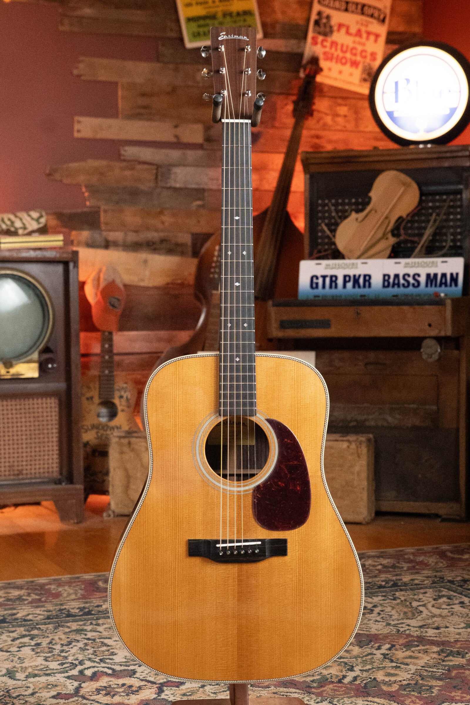 An Eastman Guitars E20D-TC Thermo-Cured Adirondack/Rosewood Dreadnought Acoustic Guitar (#8128) with a natural wood finish stands upright in a cozy, vintage-style room with wood-paneled walls, posters, and music gear.