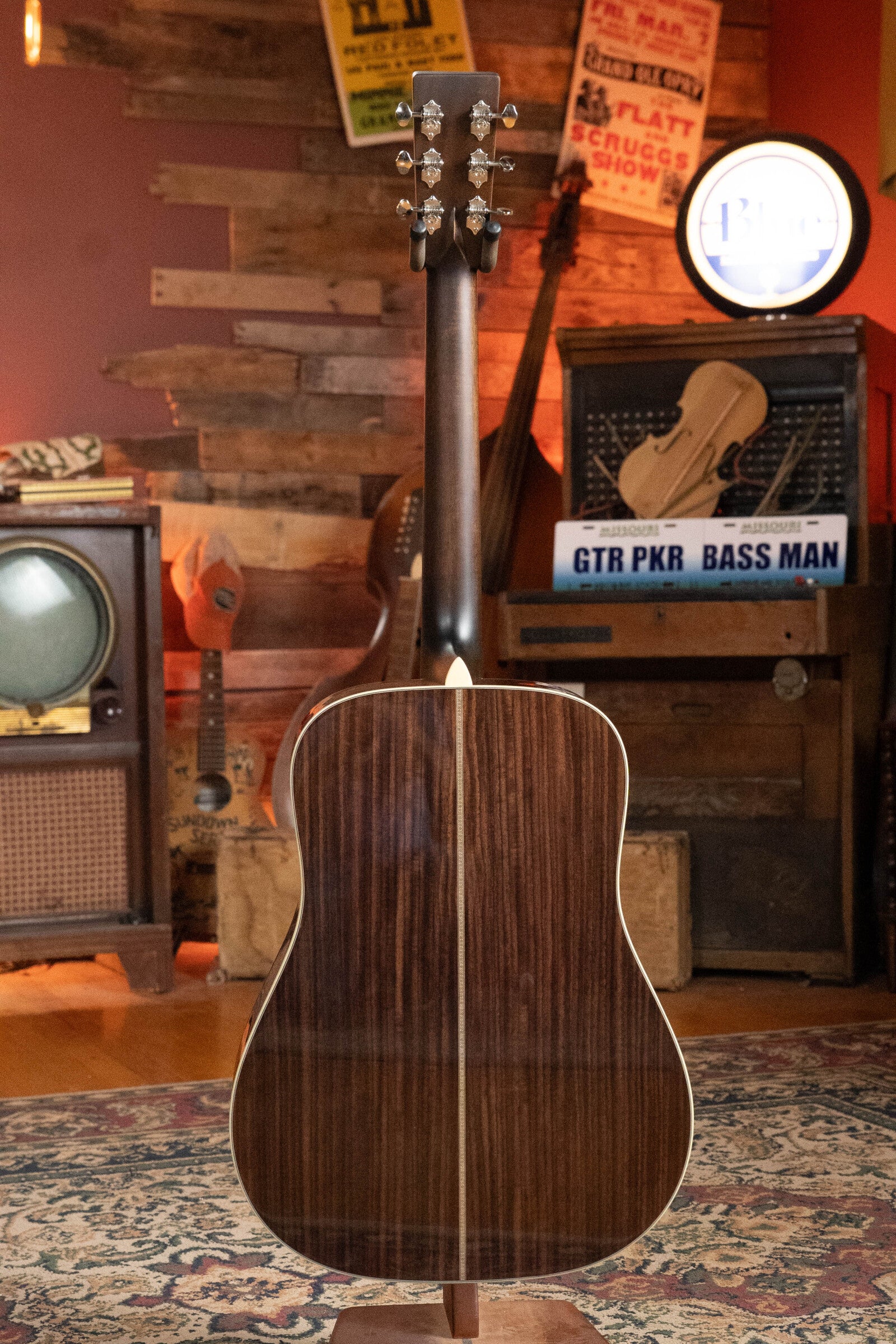 An Eastman Guitars E20D-TC Thermo-Cured Adirondack/Rosewood Dreadnought Acoustic Guitar #8128, featuring a dark rosewood back, is displayed facing away in a vintage room with retro signs, an old TV, and music equipment in the background.