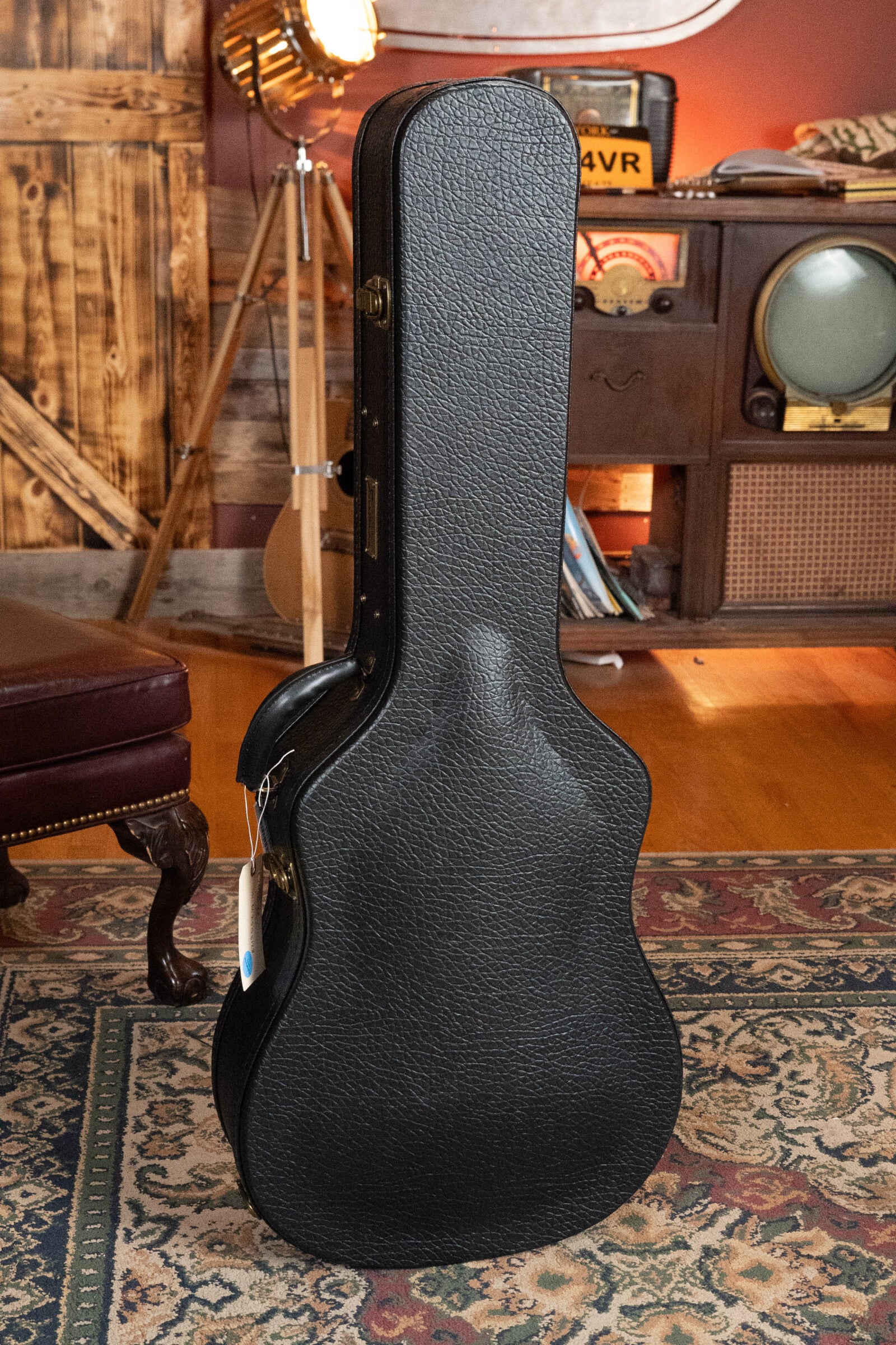 A black hard-shell case for the Eastman Guitars E20D-TC Thermo-Cured Adirondack/Rosewood Dreadnought Acoustic Guitar #8128 stands upright in a vintage-themed living room with wooden décor, an old TV, leather furniture, and a patterned rug.