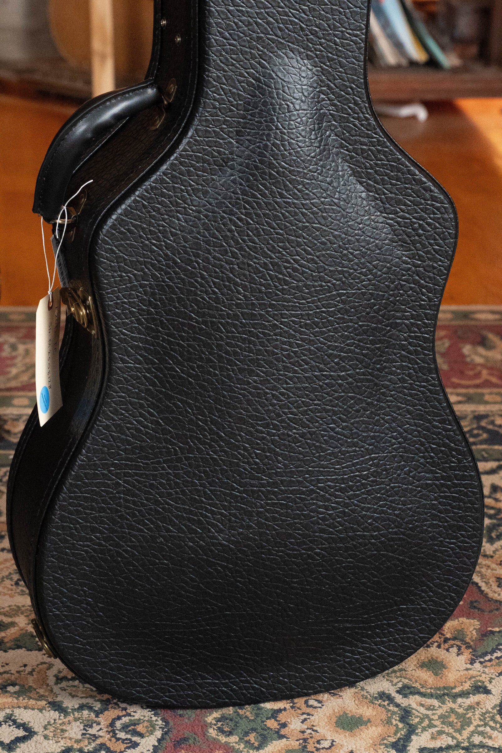 A close-up of a textured black guitar case with handle and white tag, ideal for the Eastman Guitars E20D-TC Thermo-Cured Adirondack/Rosewood Dreadnought Acoustic Guitar #8128, rests upright on patterned carpet before bookshelves.