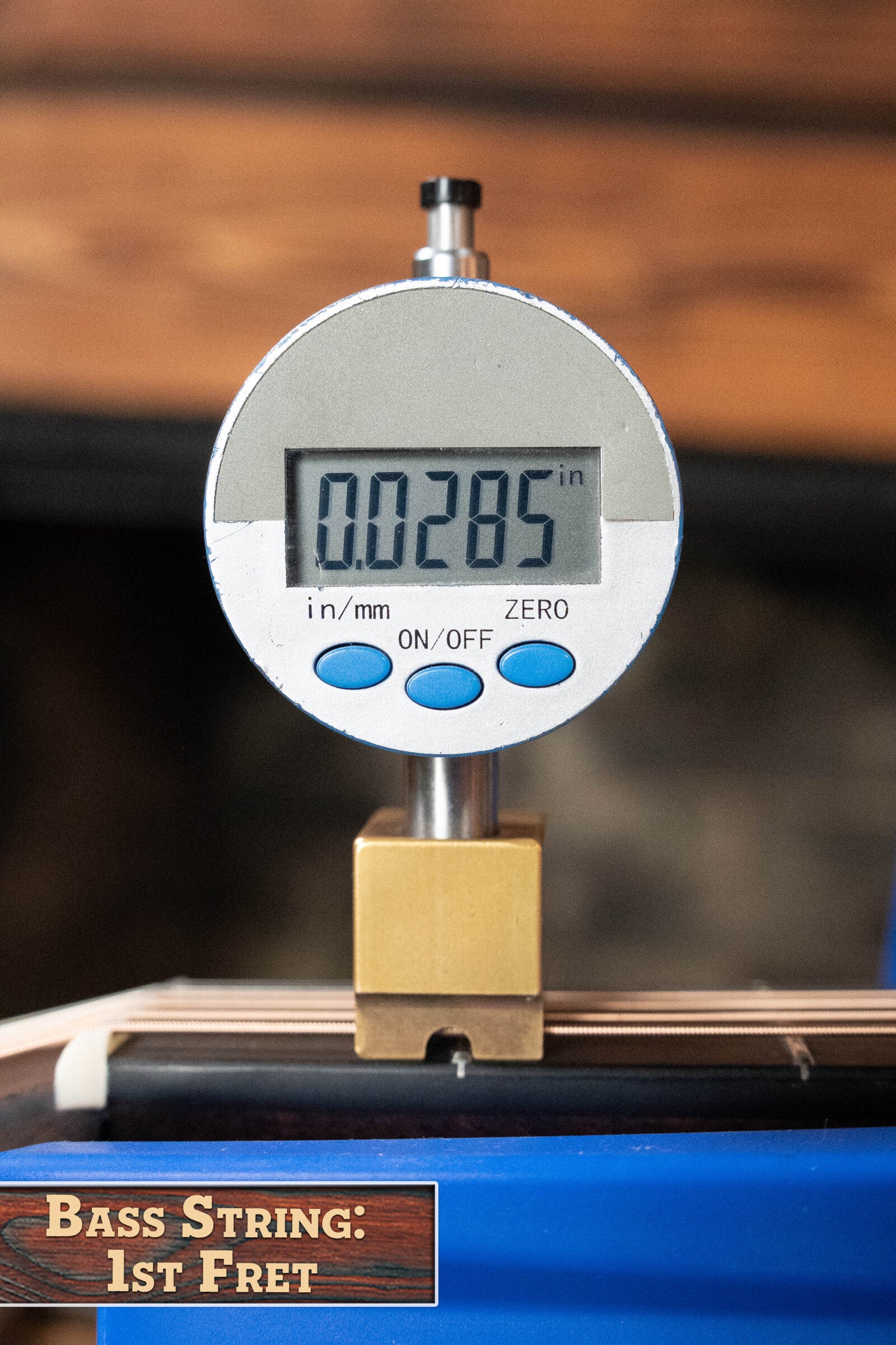 A digital gauge reads 0.0285 inches at the 1st fret of a bass string, with a display above the string on a wood background like an Eastman E20D-TC Thermo-Cured Adirondack/Rosewood Acoustic Guitar by Eastman Guitars, labeled Bass String: 1st Fret.