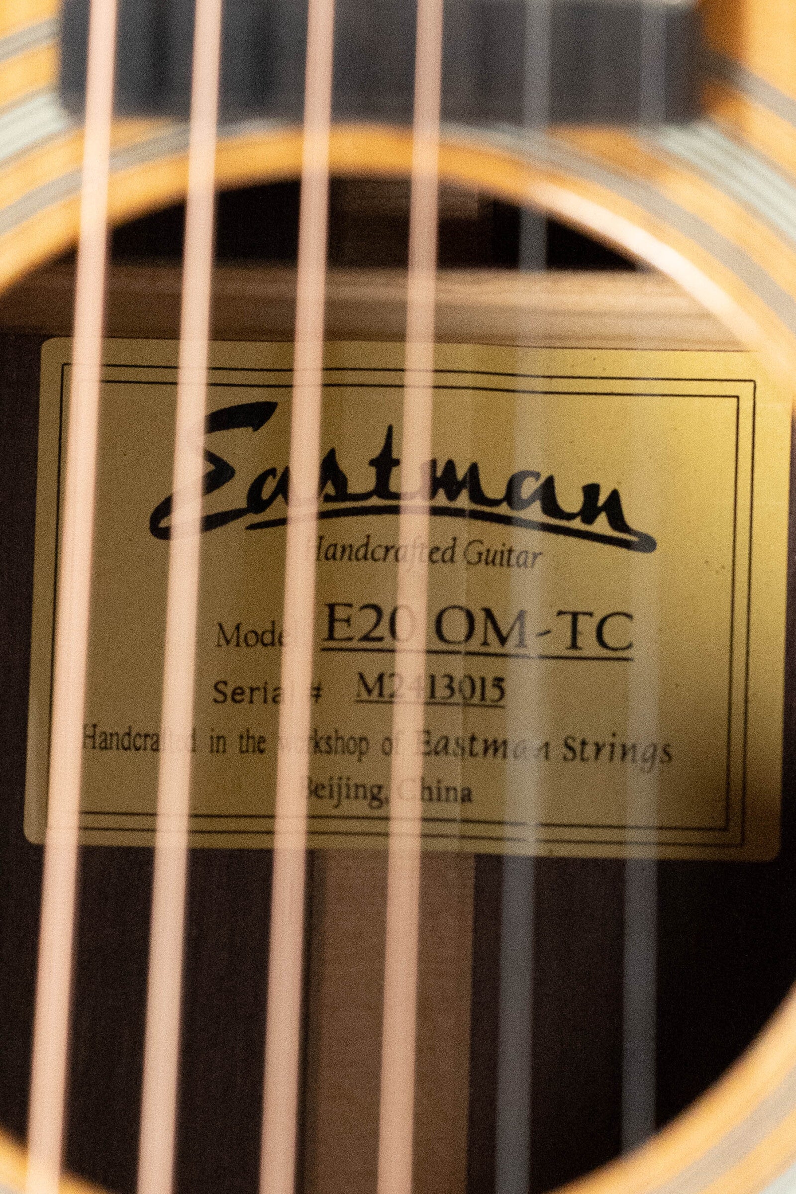 Close-up of a guitar sound hole showing a label inside that reads Eastman Guitars E20OM-TC Thermo-Cured Adirondack/Rosewood Orchestra Model Acoustic Guitar #3015, handcrafted in Beijing, China.