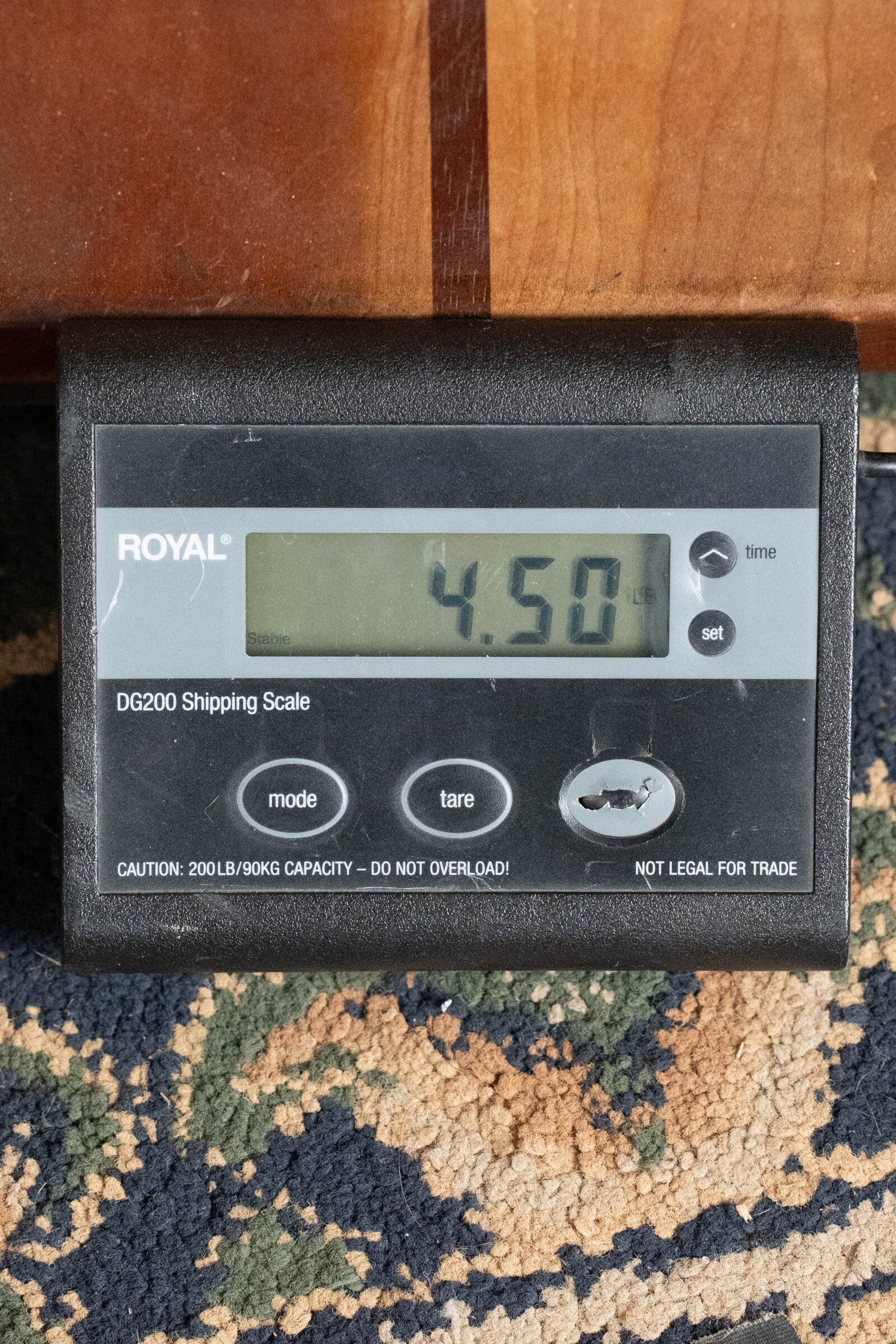 A digital shipping scale on a patterned carpet reads 4.50 lb as it weighs an Eastman Guitars E20OM-TC Thermo-Cured Adirondack/Rosewood Orchestra Model Acoustic Guitar #3015, with the black scale partially under a wooden surface.