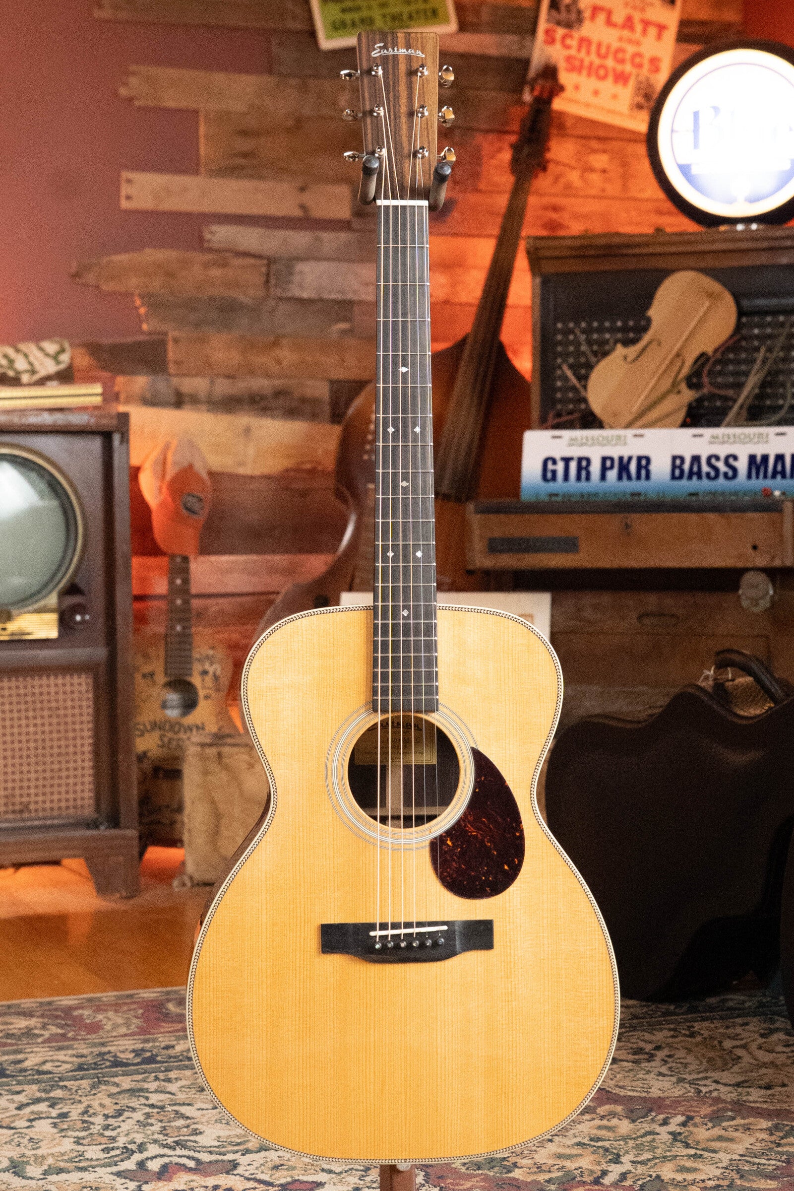 An Eastman Guitars E20OM-TC Thermo-Cured Adirondack/Rosewood Orchestra Model Acoustic Guitar #3015 with a natural finish stands on a patterned rug, set against a rustic wooden wall with vintage TV and music gear in the background.