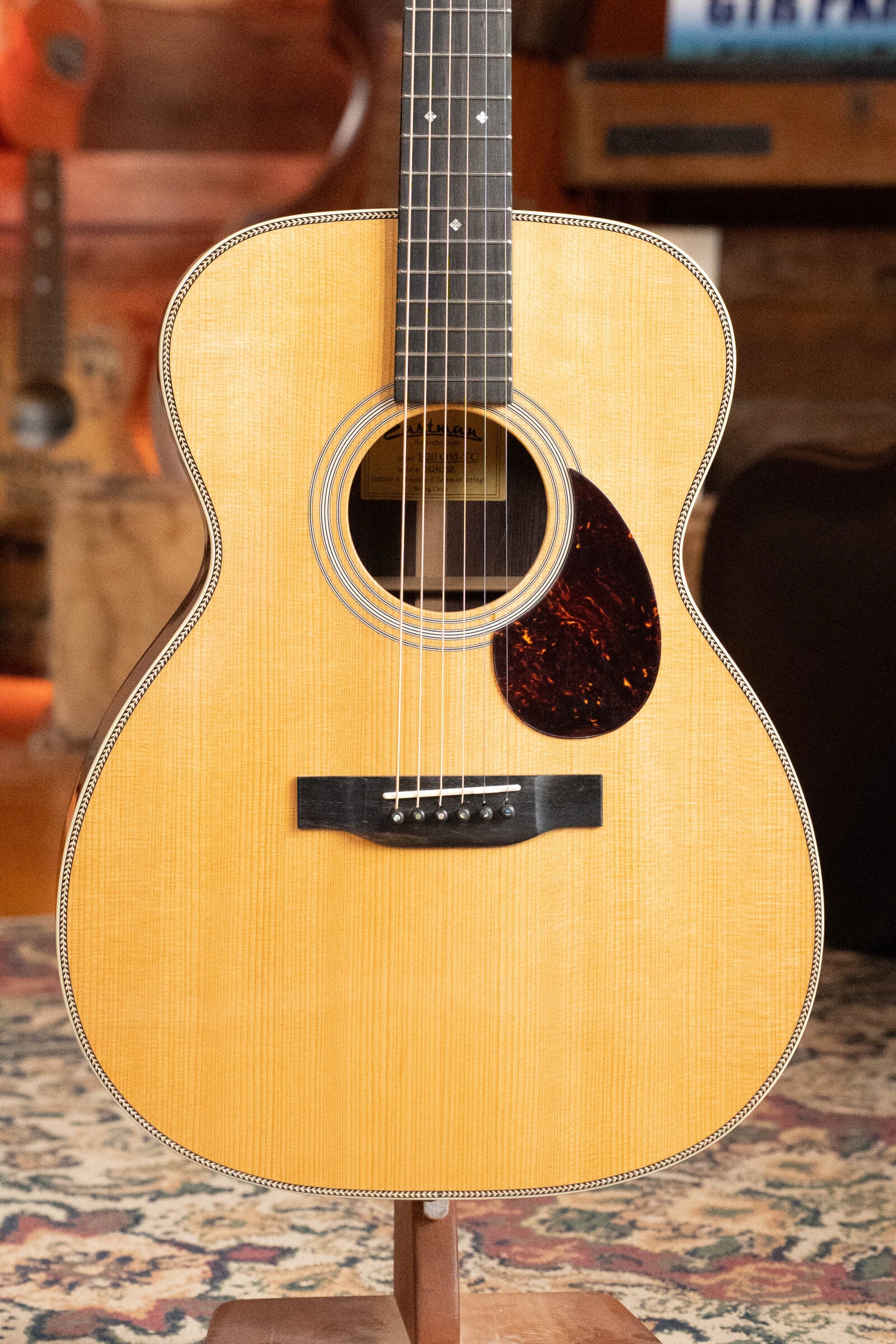 An Eastman Guitars E20OM-TC Thermo-Cured Adirondack/Rosewood Orchestra Model Acoustic Guitar #3015 is displayed indoors on a stand, featuring a light wood body, dark pickguard, and rosette, with other guitars and a rug in the background.
