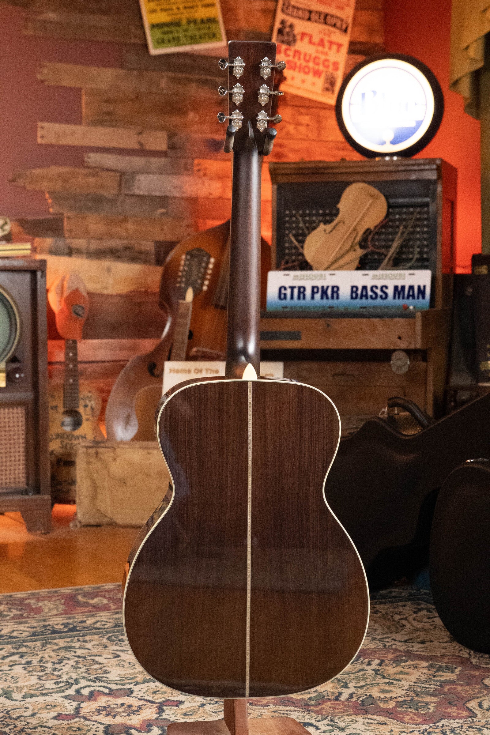 An Eastman E20OM-TC Thermo-Cured Adirondack/Rosewood Orchestra Model Acoustic Guitar (#3015) by Eastman Guitars stands with its back to the camera in a cozy, vintage room with musical decor, wood walls, posters, and a patterned rug.