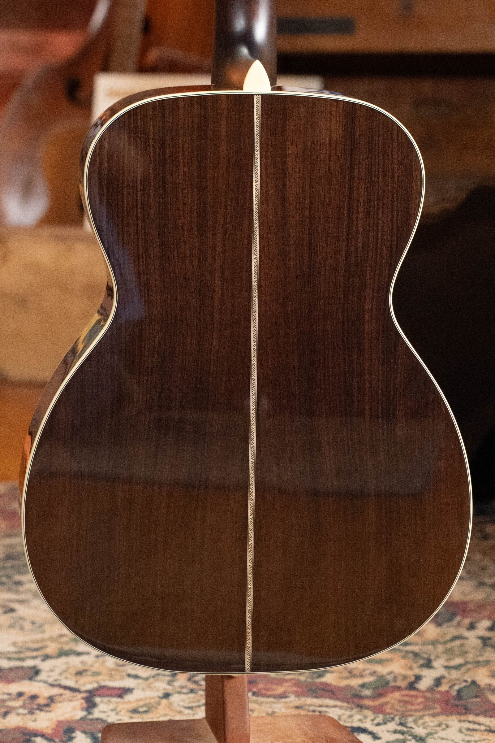 The image shows the back of an Eastman Guitars E20OM-TC Thermo-Cured Adirondack/Rosewood Orchestra Model Acoustic Guitar #3015 with a glossy dark rosewood finish, thin light center inlay, and cream binding, standing upright on a patterned rug.