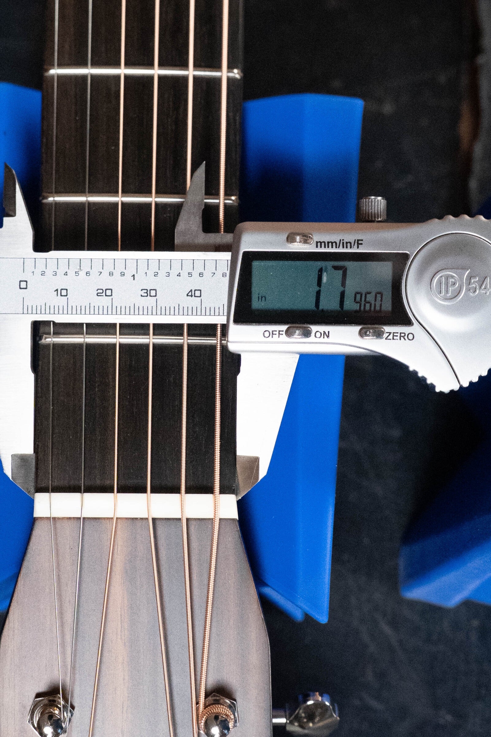 An Eastman E20OM-TC guitar by Eastman Guitars has its rosewood neck width measured at 1.7 inches with a digital caliper; the headstock holds six strings and blue supports appear on each side.