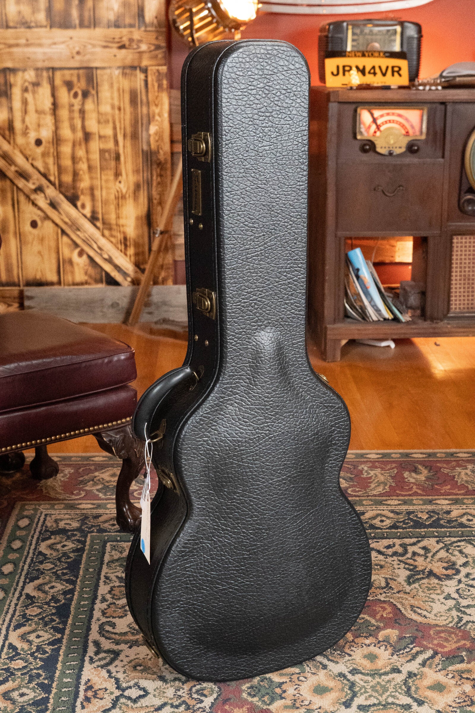 A black hard-shell guitar case stands upright on a patterned rug in a cozy room, ready to protect an Eastman Guitars E20OM-TC Thermo-Cured Adirondack/Rosewood Orchestra Model Acoustic Guitar #3015. A rustic door adds charm to the space.