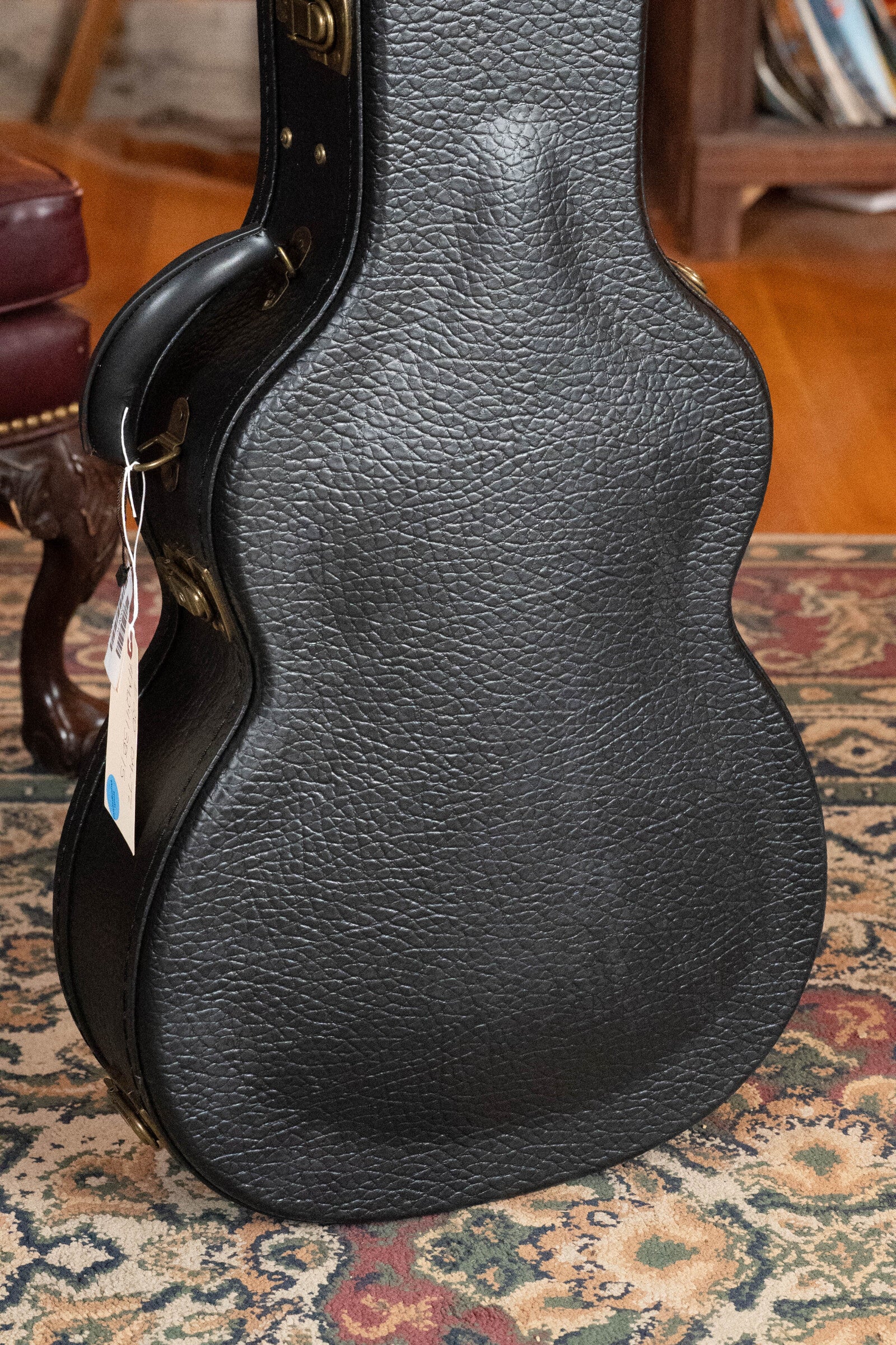 A black textured hard case for the Eastman Guitars E20OM-TC Thermo-Cured Adirondack/Rosewood Orchestra Model Acoustic Guitar (#3015) stands upright on a patterned rug, with a chair and wooden shelf in the background.